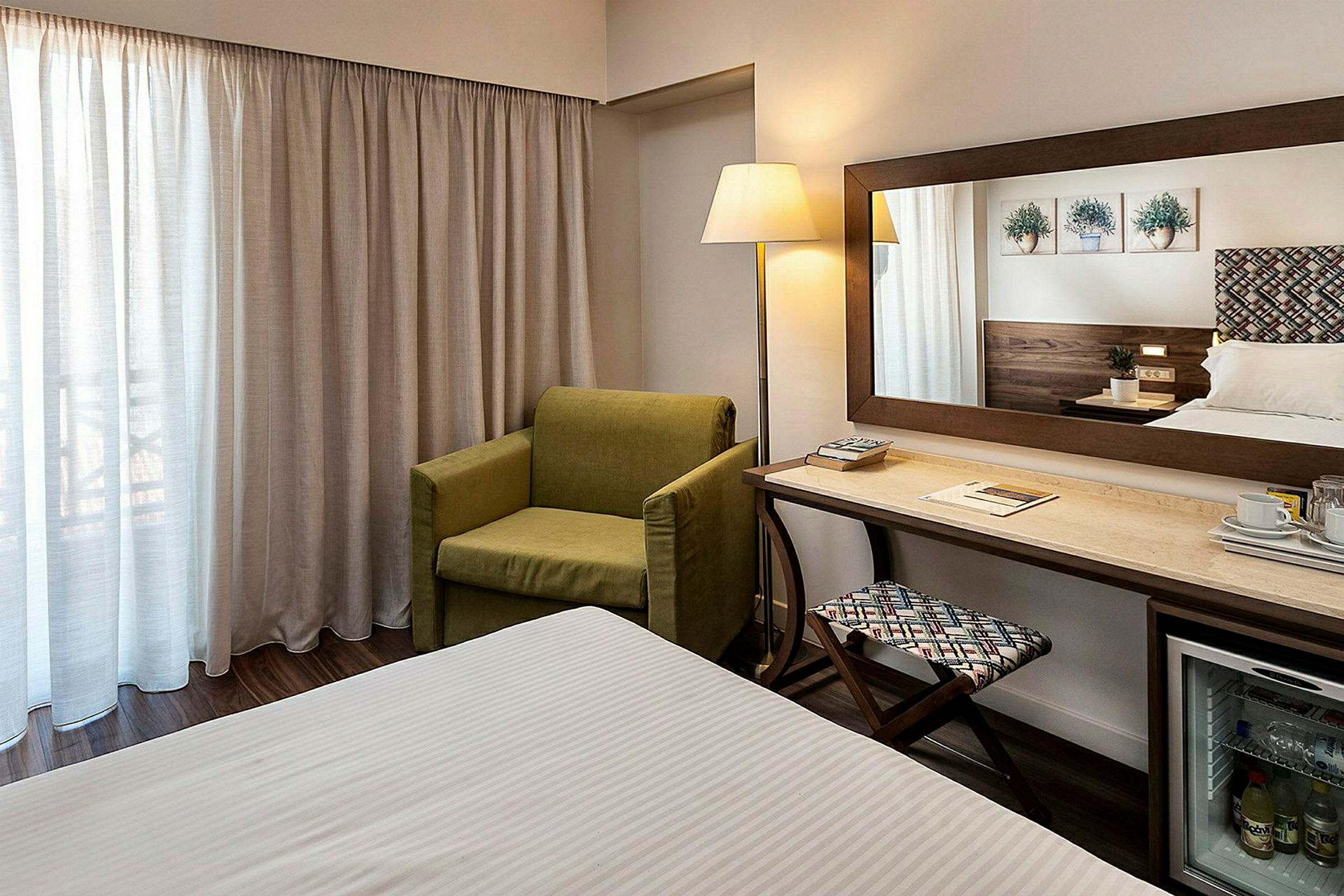 Hotelværelse med grøn lænestol, skrivebord, spejl og seng. (AI-genereret tekst)