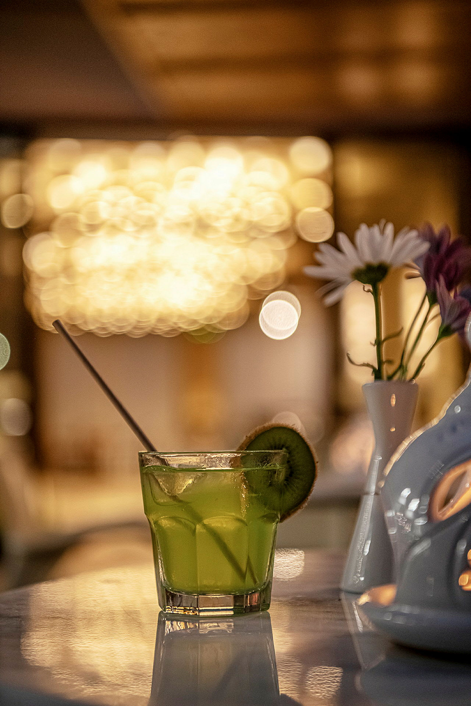Et glas med en grøn drink pyntet med en kiwiskive på en marmorbord med en vase med blomster i baggrunden. (AI-genereret tekst)
