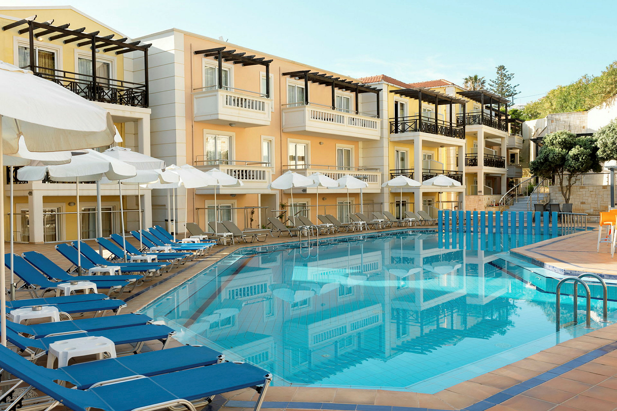 Hotellets facade i pastelfarver med altaner, foran en blå swimmingpool omgivet af solsenge og parasoller. (AI-genereret tekst)