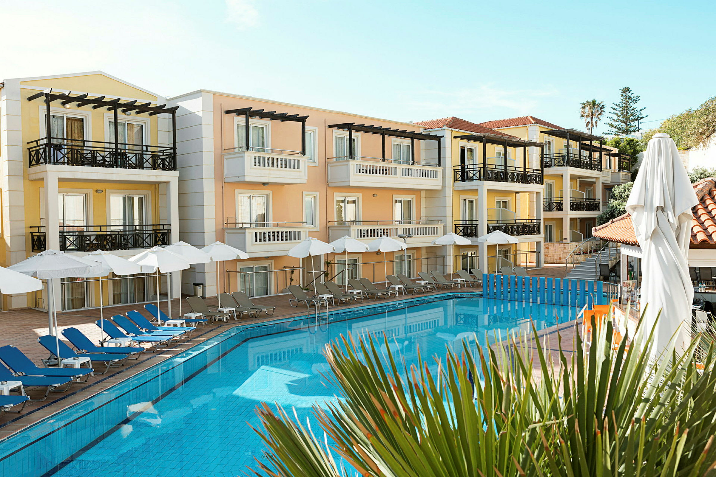 Hotelbygninger med altaner omkring en swimmingpool samt poolområde med liggestole og parasoller. (AI-genereret tekst)
