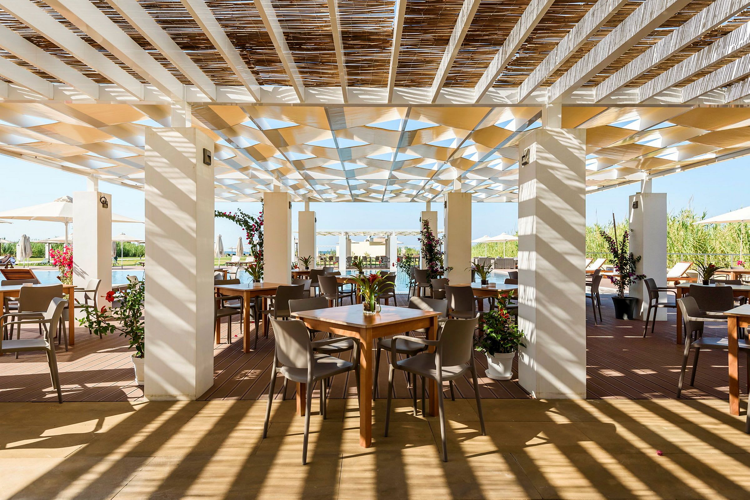 En udendørs restaurant med borde og stole under en skyggefuld pergola, omgivet af grønne omgivelser. (AI-genereret tekst)