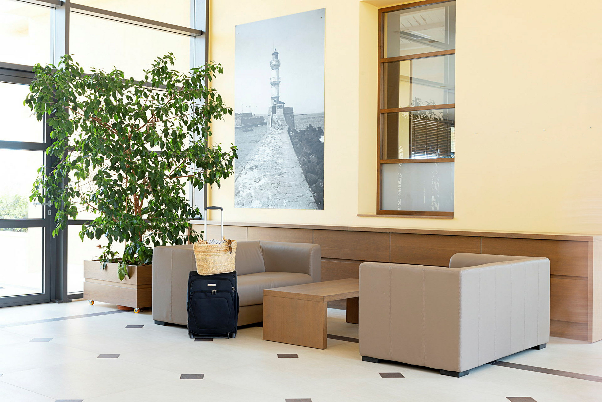 En receptionslounge med beige lænestole, et bord, en stor grøn plante samt en sort kuffert med en kurv ovenpå. (AI-genereret tekst)