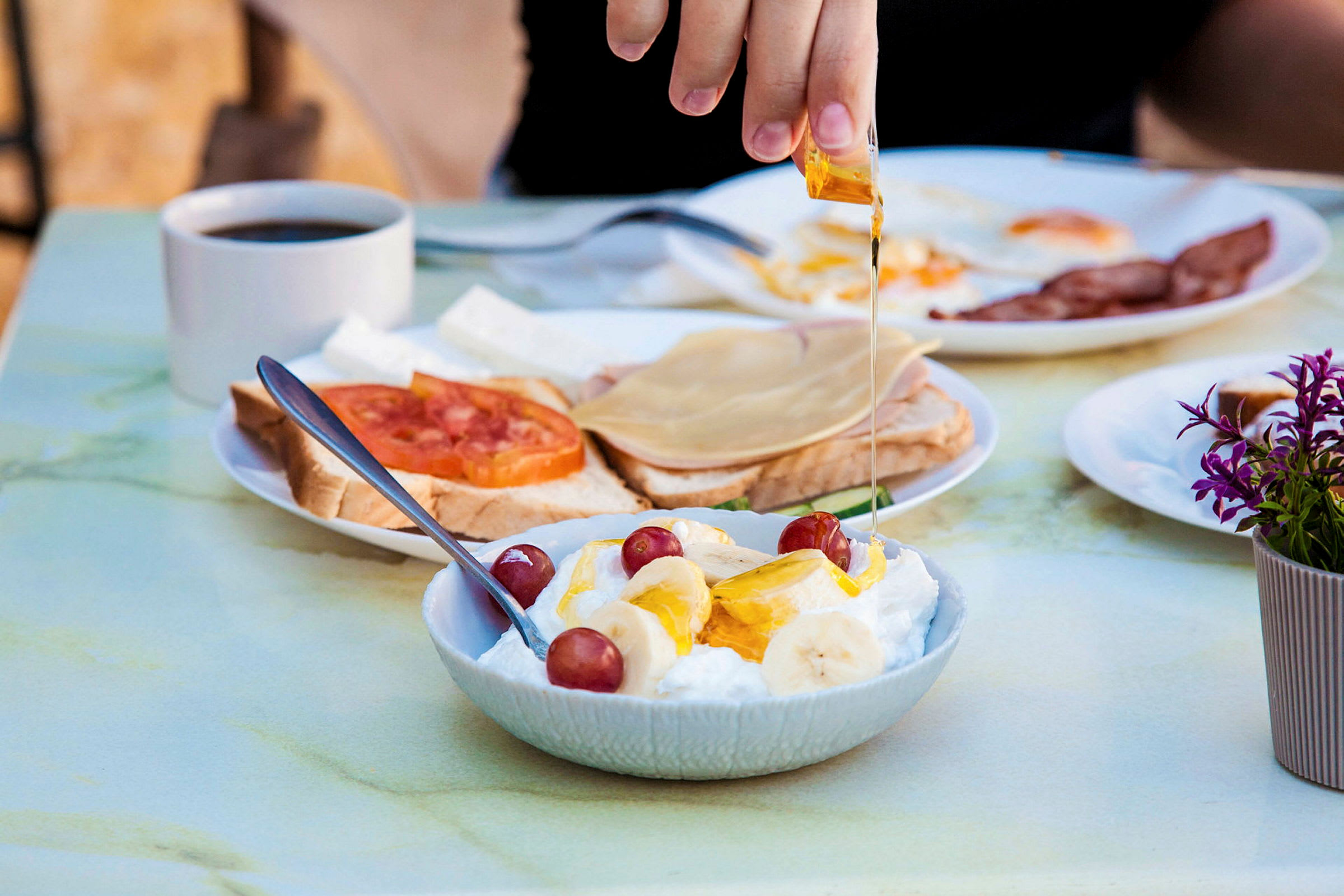 En hånd hælder honning over en skål med yoghurt, banan og druer. I baggrunden er der en kaffekop og tallerkener med brød og pålæg. (AI-genereret tekst)