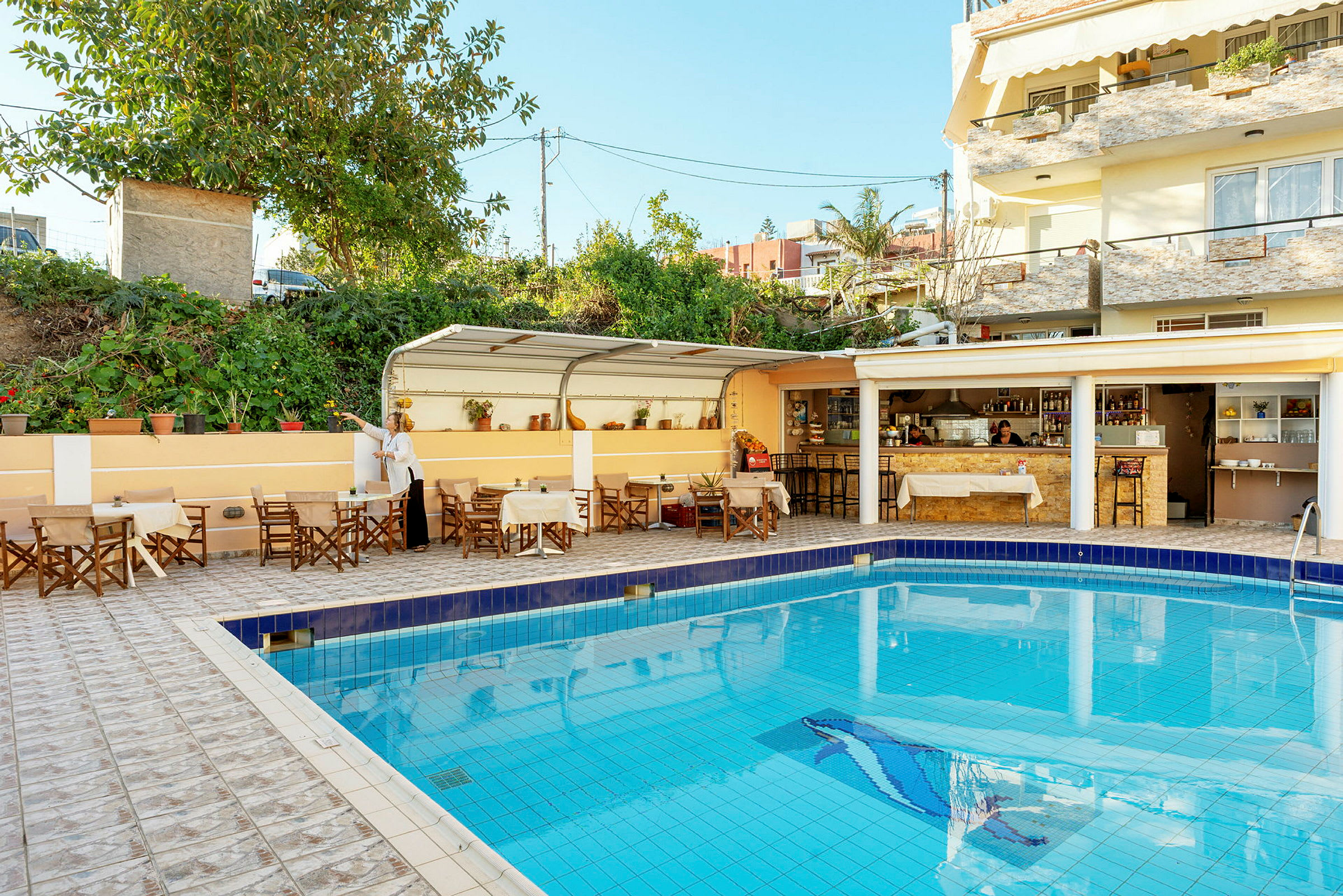 Udendørsområde med en pool omgivet af borde og stole. I baggrunden en overdækket café- eller barservering. (AI-genereret tekst)