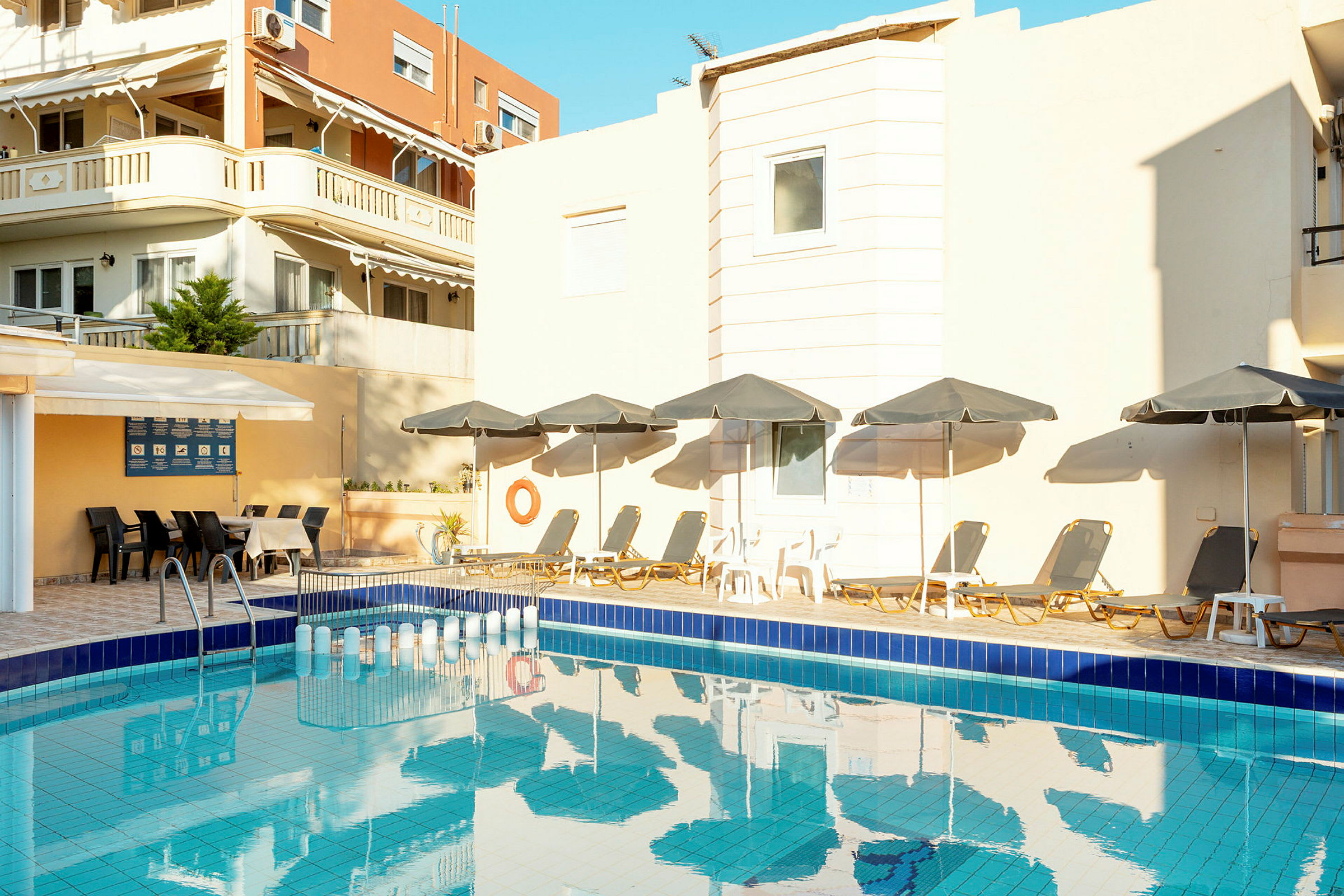 En swimmingpool omgivet af liggestole med parasoller og en bygning i baggrunden. (AI-genereret tekst)