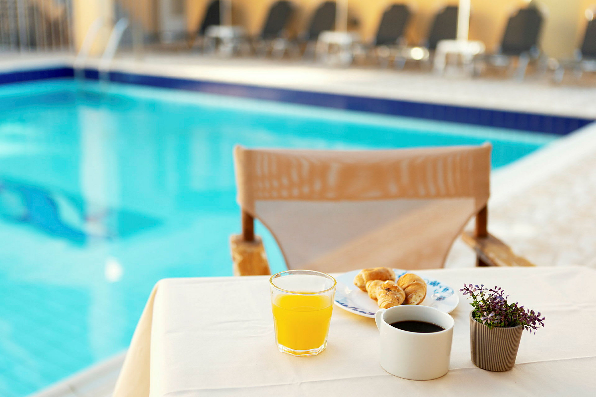 En borddug med en morgenmad opstilling ved en pool, inkluderer juice, kaffe, croissanter og en lille plante. (AI-genereret tekst)
