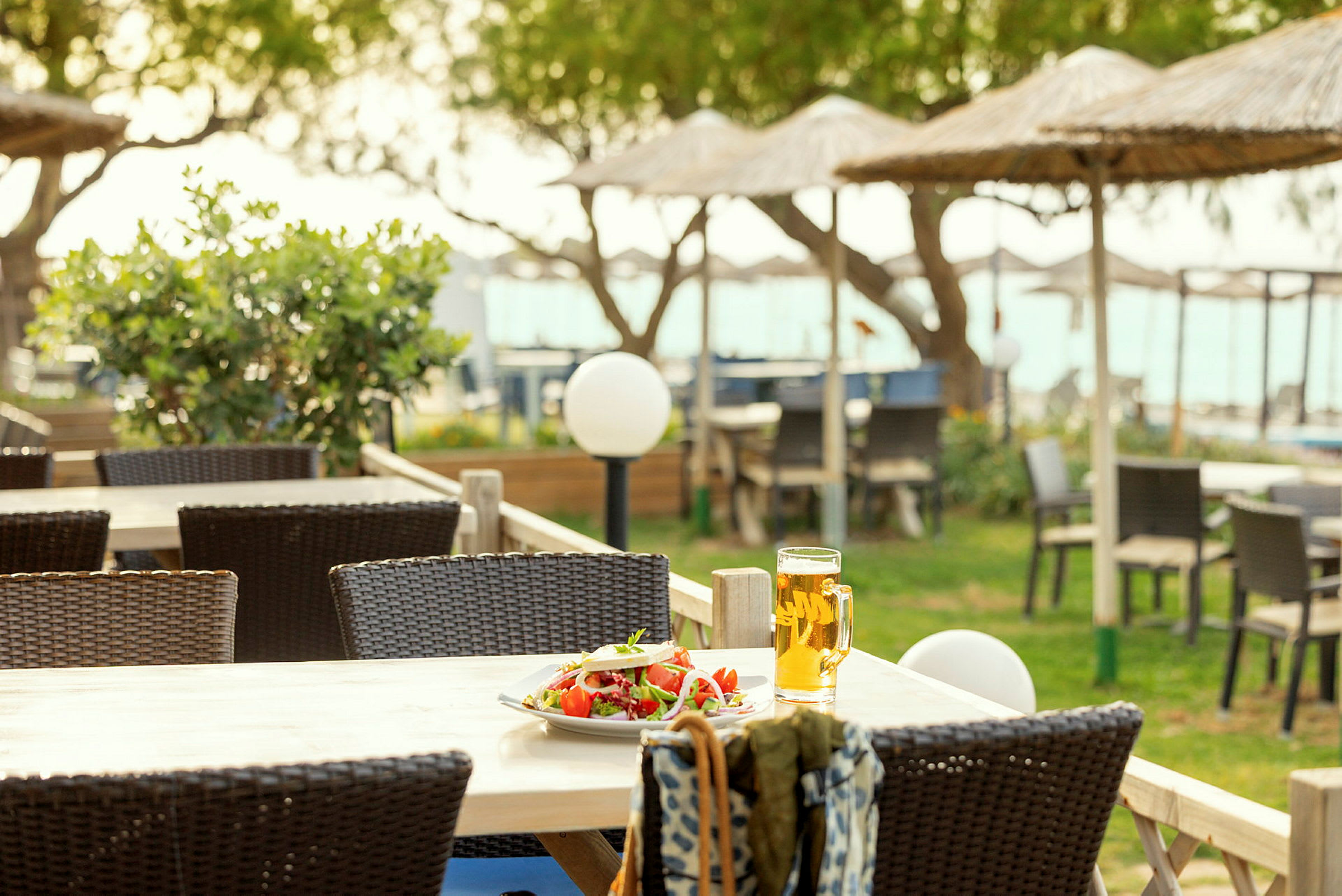 Et bord med en tallerken salat og et glas øl på en terrasse med havemøbler og strandparasoller i baggrunden. (AI-genereret tekst)