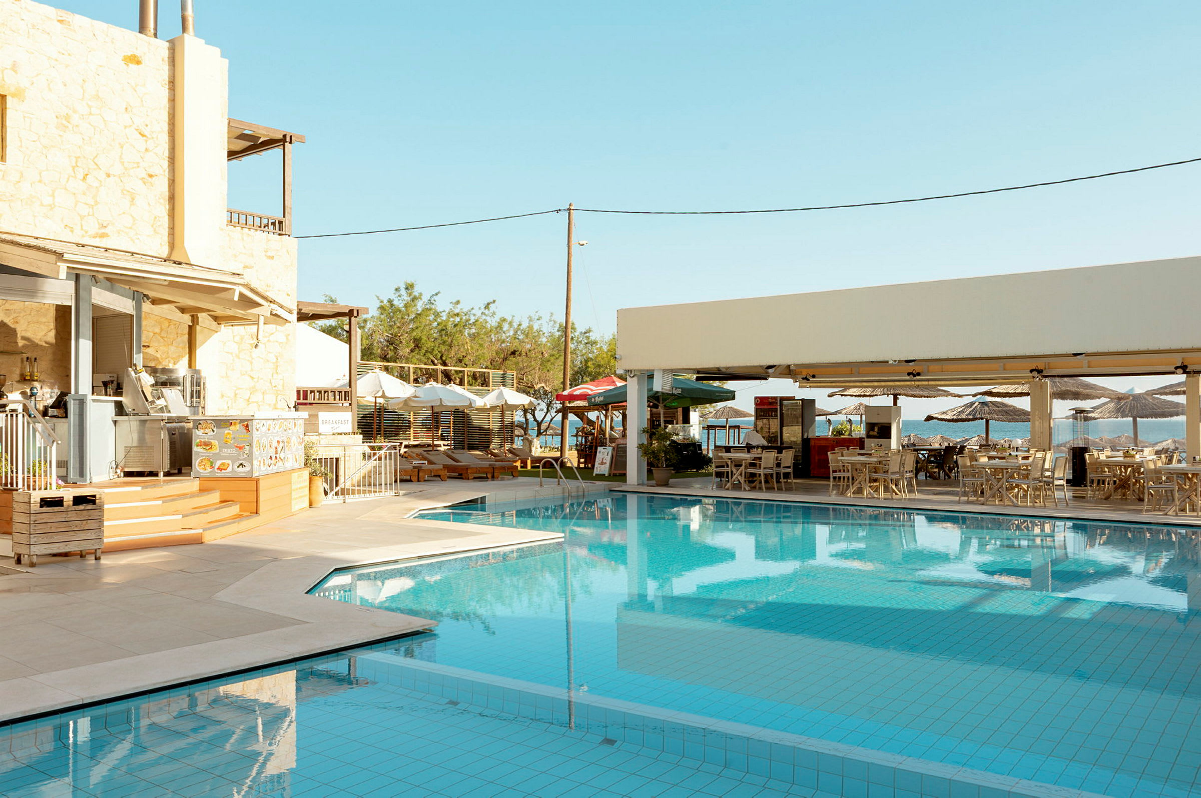 Billede af en udendørs swimmingpool ved siden af restauranter og siddepladser, omgivet af liggestole og parasoller. (AI-genereret tekst)