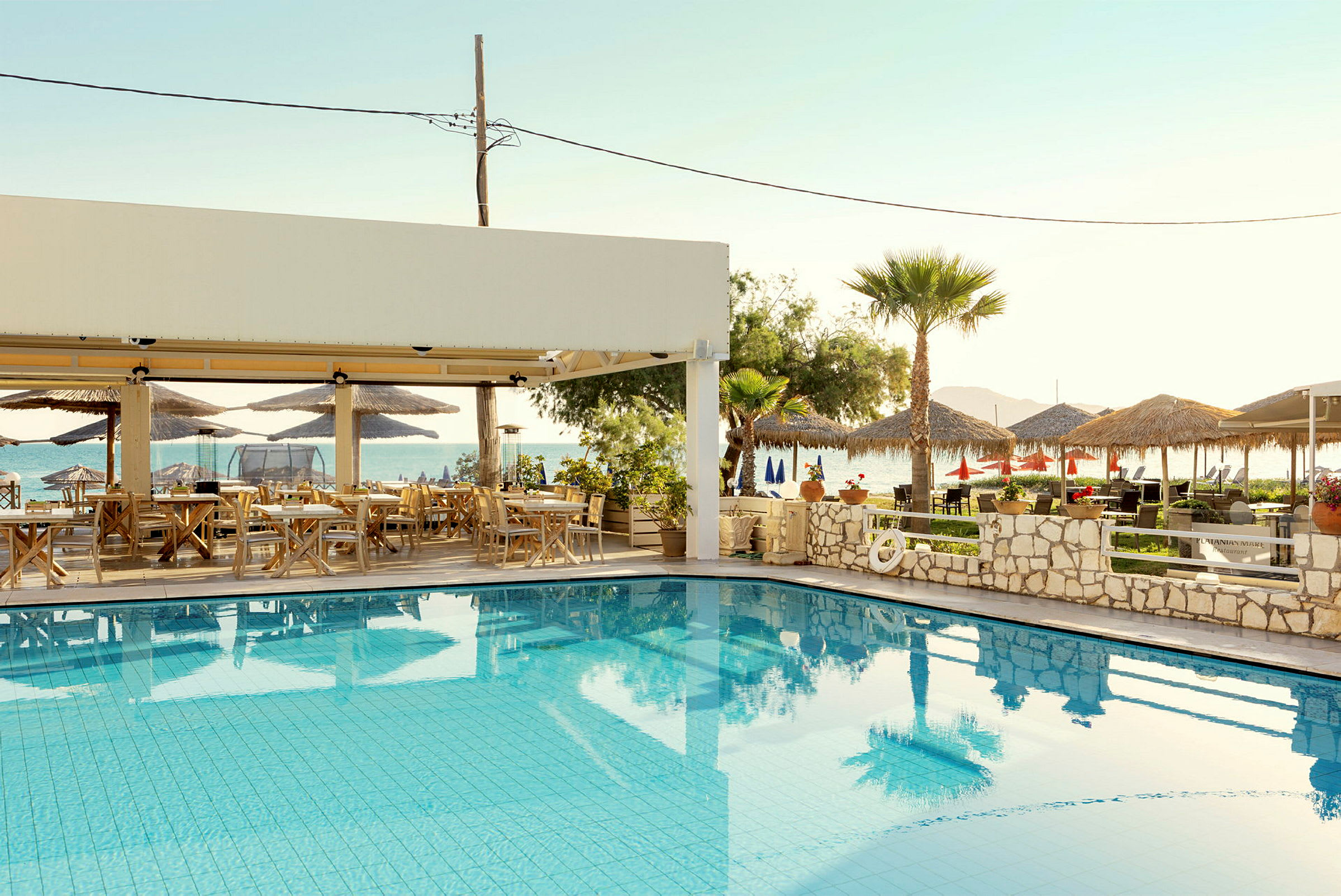 En udendørs pool med siddepladser under en skyggefuld pergola og udsigt over havet. (AI-genereret tekst)