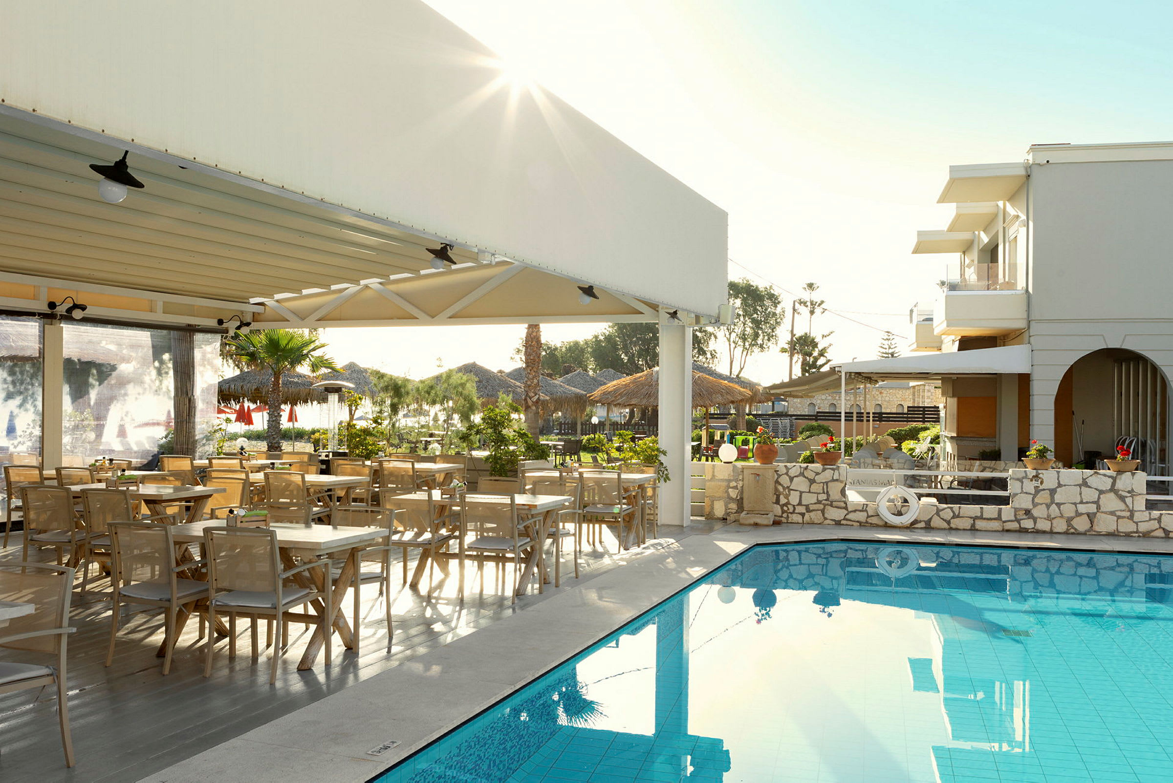 Udendørs område ved en restaurant med siddepladser og en pool. Bygninger og beplantning ses i baggrunden. (AI-genereret tekst)