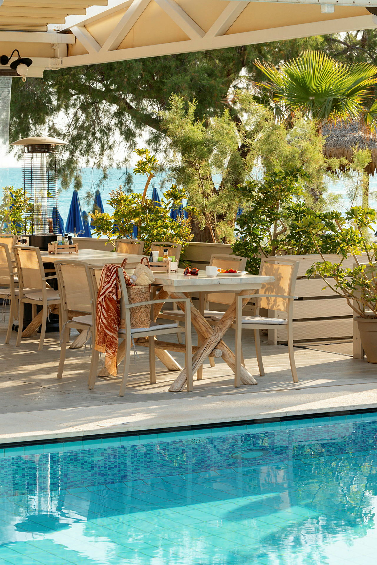 En udendørs terrasse med borde og stole tæt på en pool, omgivet af beplantning og med havet i baggrunden. (AI-genereret tekst)