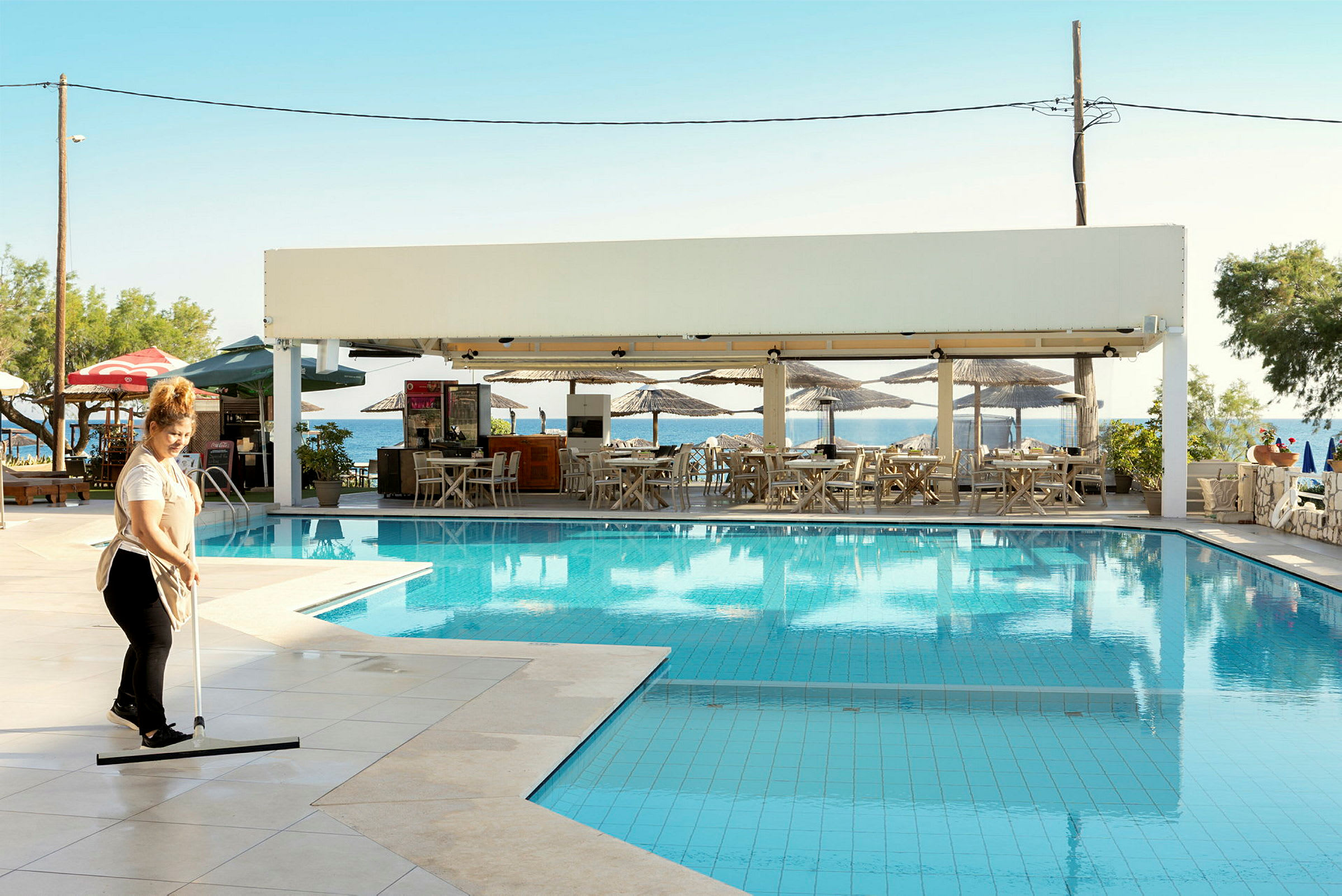 En kvinde rengør området ved en pool tæt på en udendørs restaurant med solskærme. Restauranten ligger ved havet. (AI-genereret tekst)
