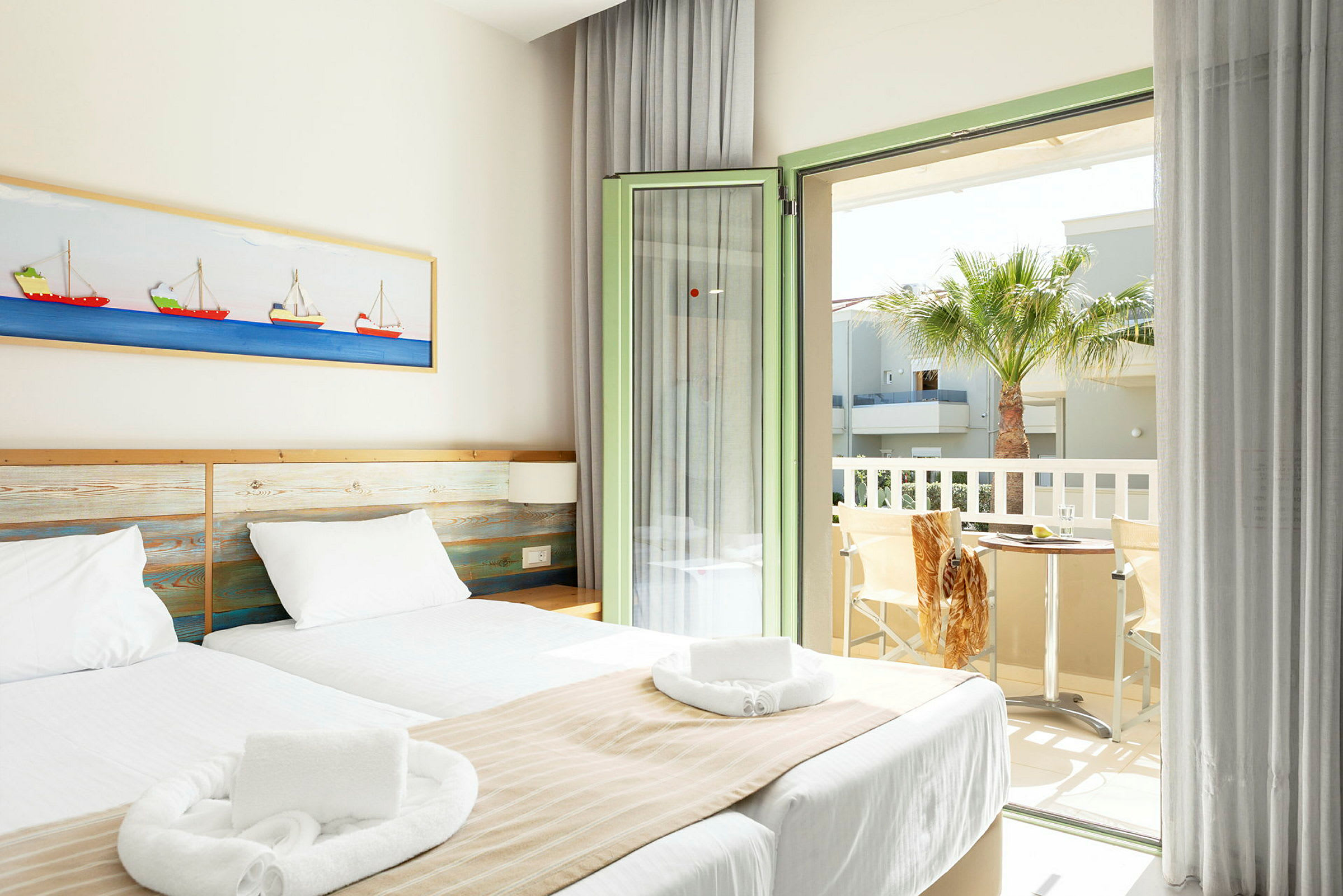 Et lyst hotelværelse med to enkeltsenge, hvidt sengetøj og beige sengetæpper. Et billede med bådmotiver hænger på væggen over sengen. Værelset har en åben altandør med udsigt til en palme og et bord med to stole. (AI-genereret tekst)
