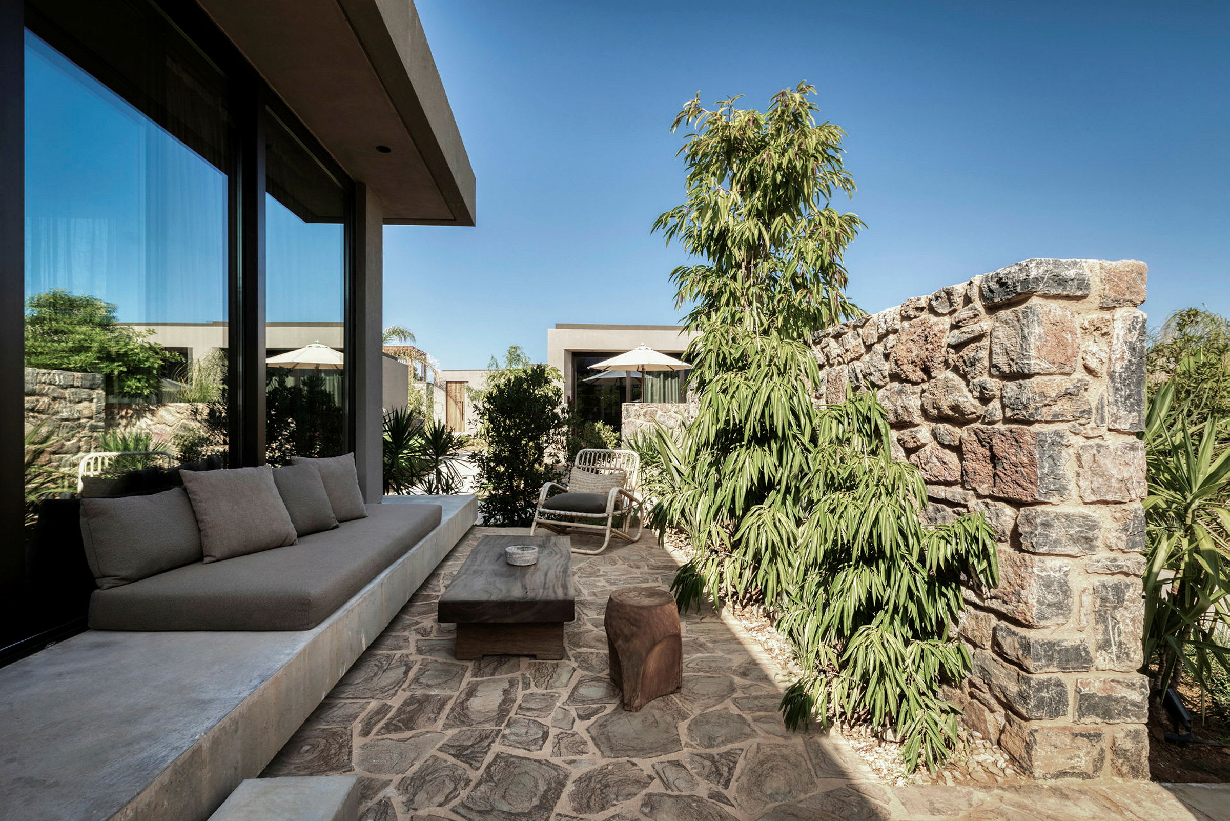 Stenbelagt terrasse med sofa, bord og stole nær et glashus, omgivet af planter og mure. (AI-genereret tekst)