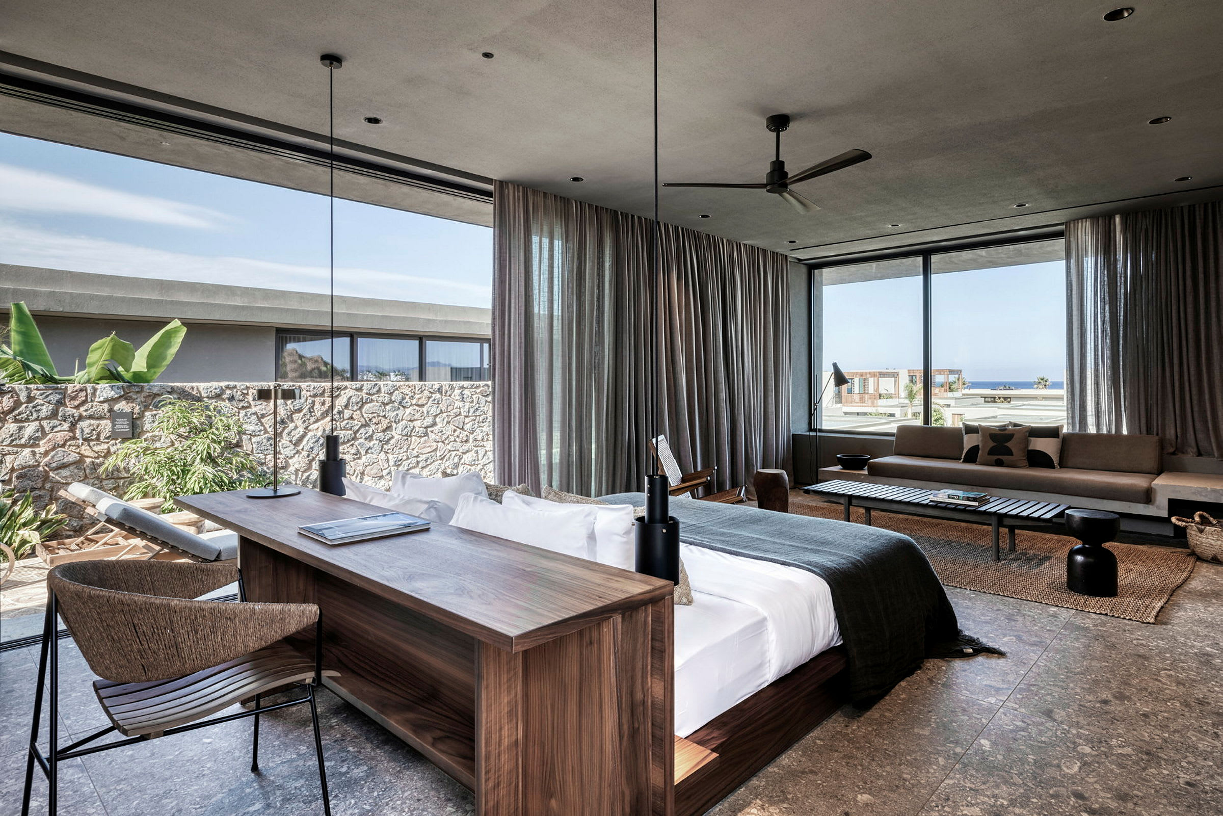 Moderne hotelsuite med stor seng, skrivebord og siddeområde. Vinduer med udsigt over havet og grønne omgivelser. (AI-genereret tekst)