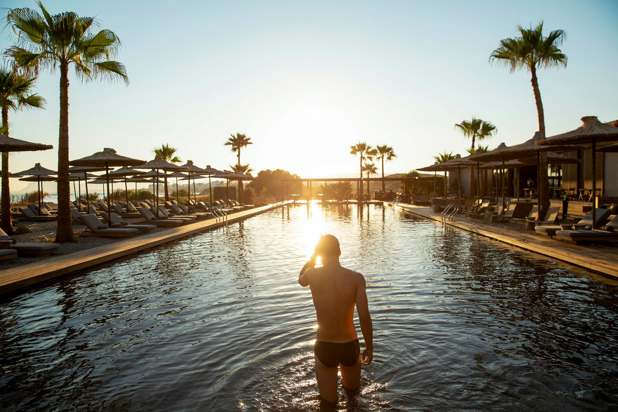 Person, der går i en pool ved solnedgang med liggestole og parasoller omkring. (AI-genereret tekst)