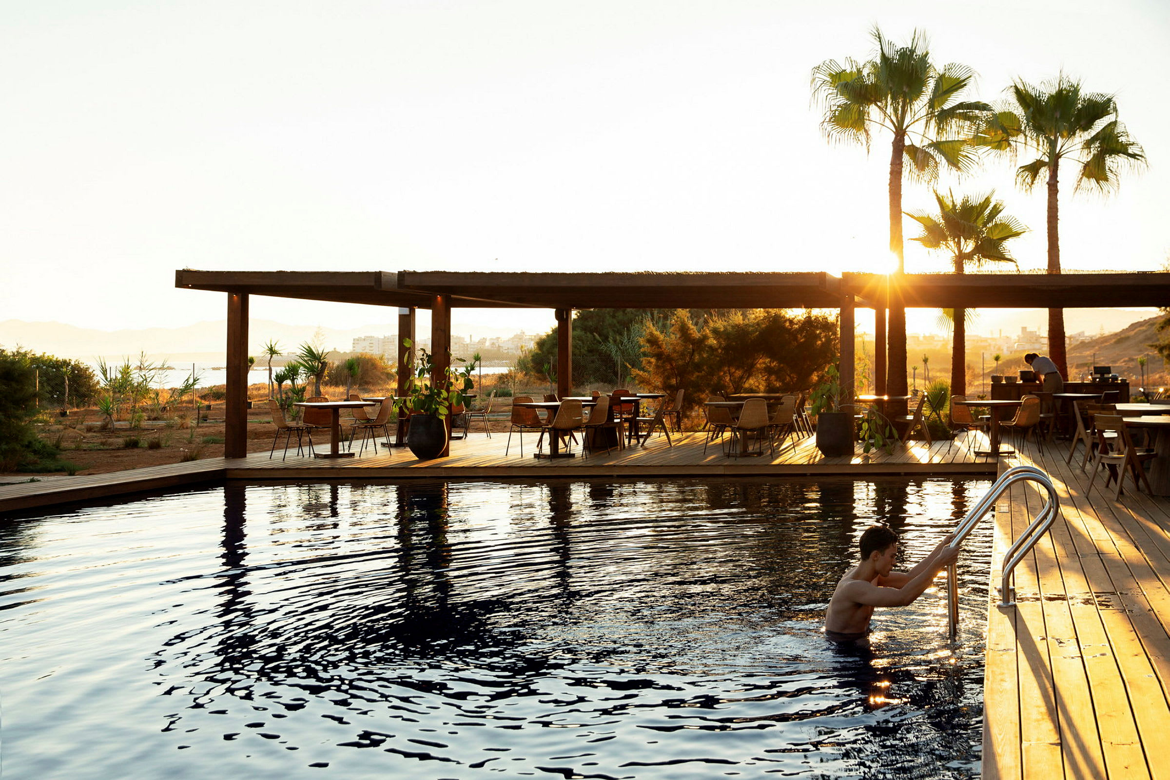 En person holder ved en poolstige ved en udendørs pool. I baggrunden er der en terrasse med borde og stole samt palmer i solnedgang. (AI-genereret tekst)