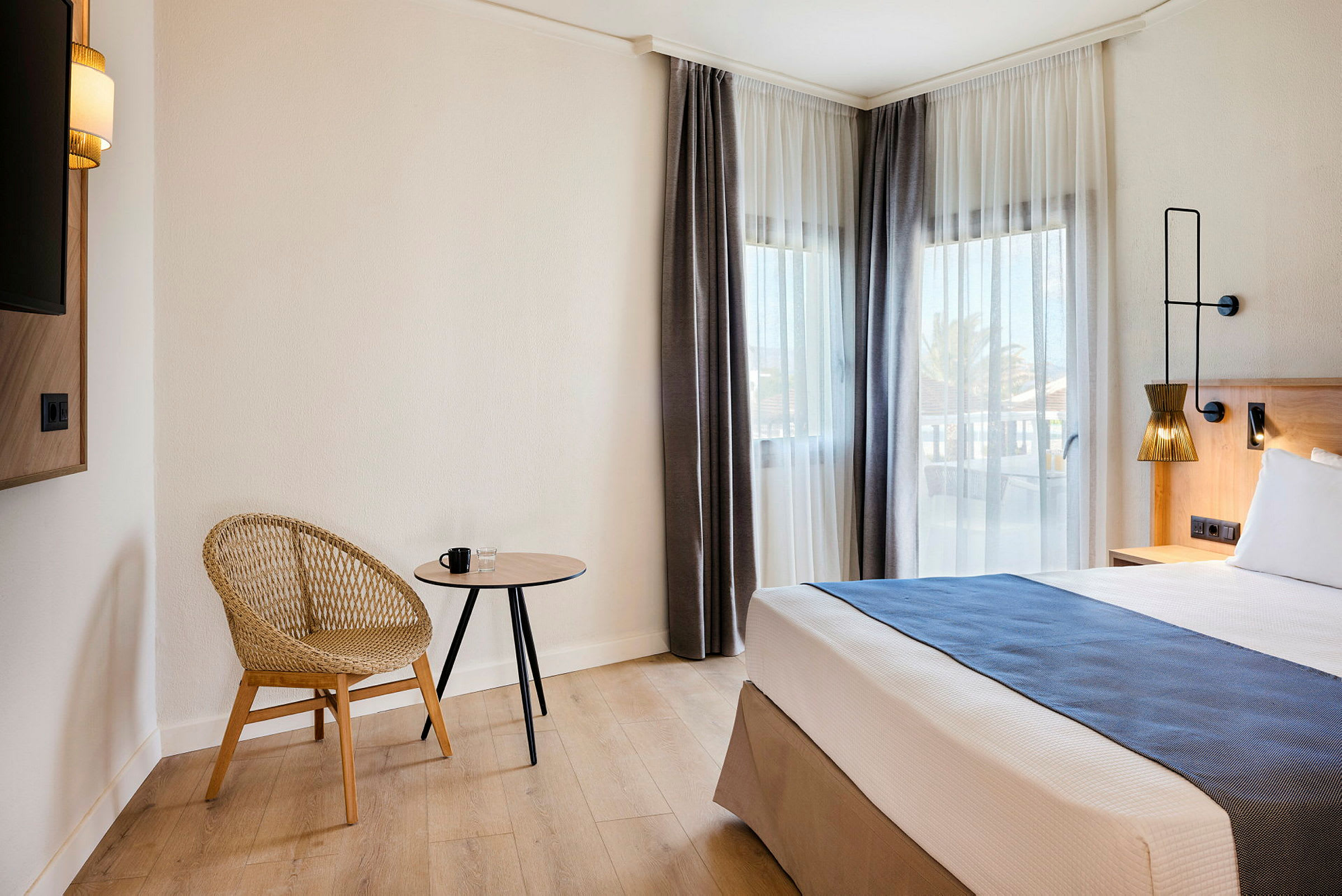 Et hotelværelse med en moderne stol, et lille bord og en seng ved vinduer med lyse gardiner. (AI-genereret tekst)