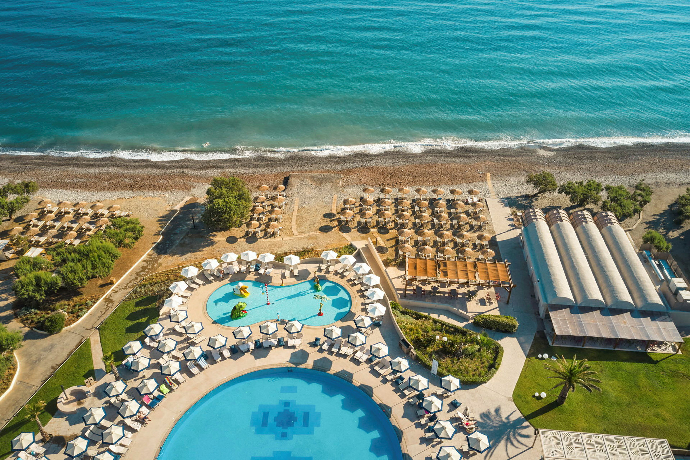 Luftfoto af hotelområde med pools, parasoller og stranden ved havet. (AI-genereret tekst)