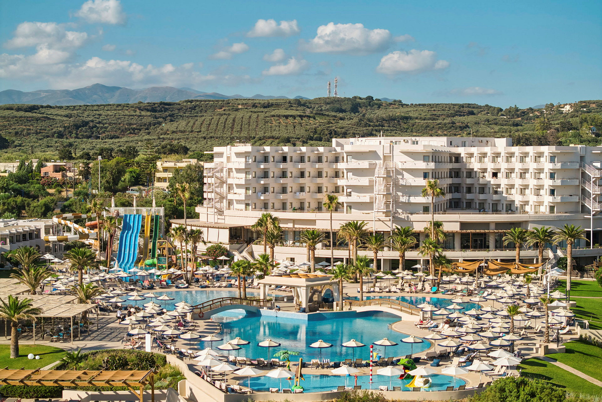 Resort med stort poolområde og vandrutsjebaner, omgivet af grønt landskab. (AI-genereret tekst)