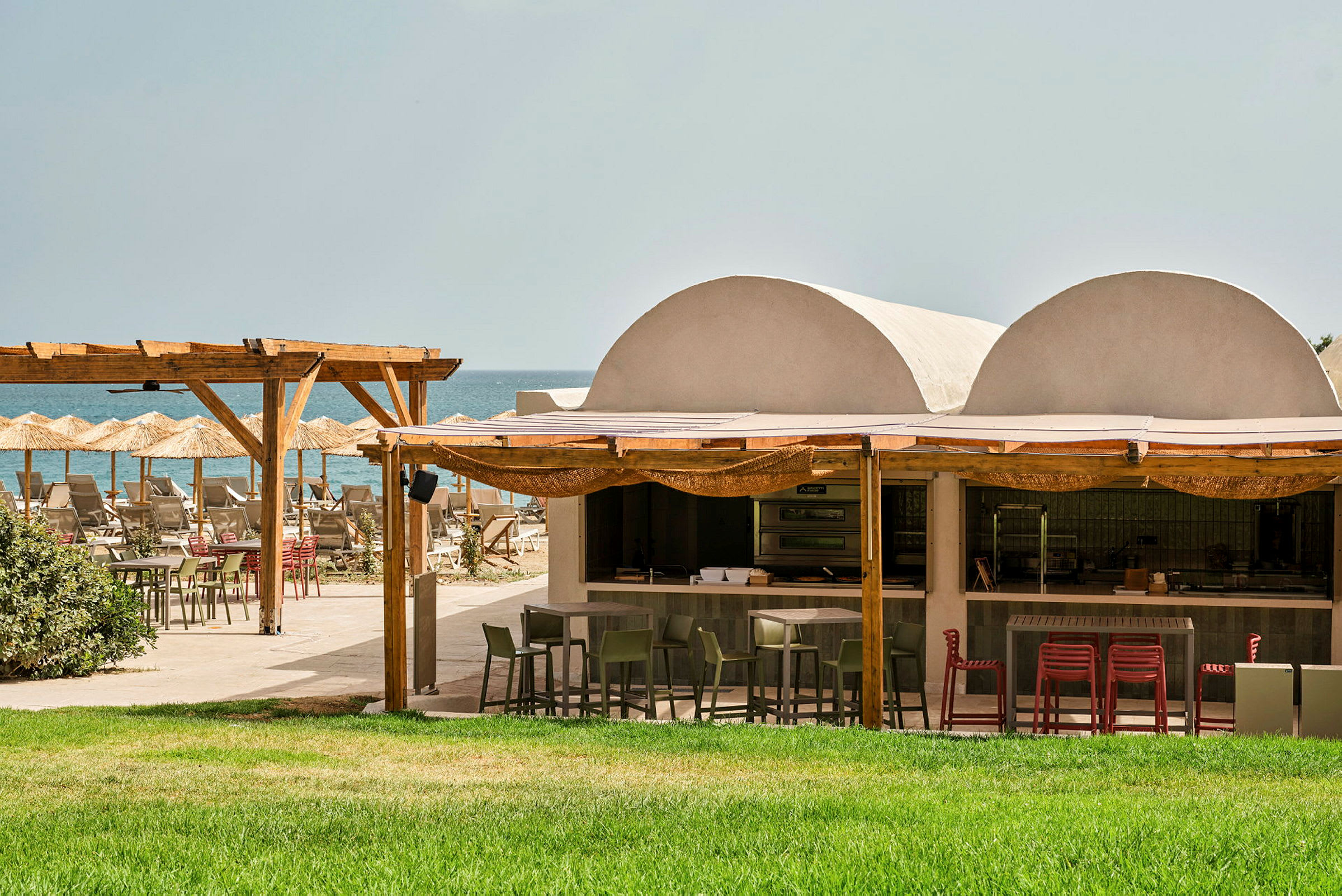 Udendørs restaurant ved stranden med grøn græsplæne, træpergola og parasoller. (AI-genereret tekst)
