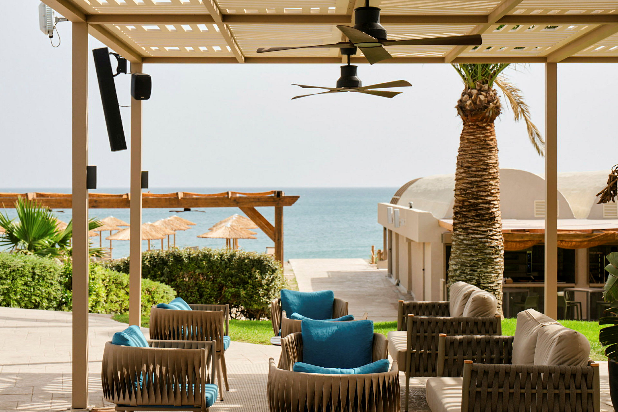 Havemøbler under en pergola med udsigt til en sandstrand og havet i baggrunden. (AI-genereret tekst)