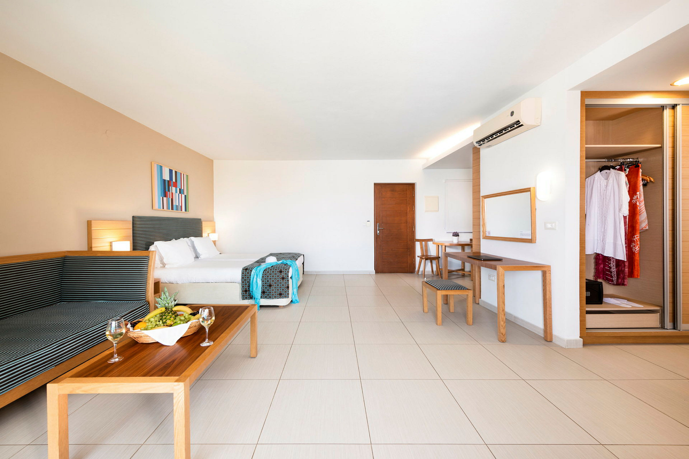 Et rummeligt hotelværelse med dobbeltseng, siddeområde, spisebord, spejl og garderobe. En frugtskål og vinglas står på sofabordet. (AI-genereret tekst)