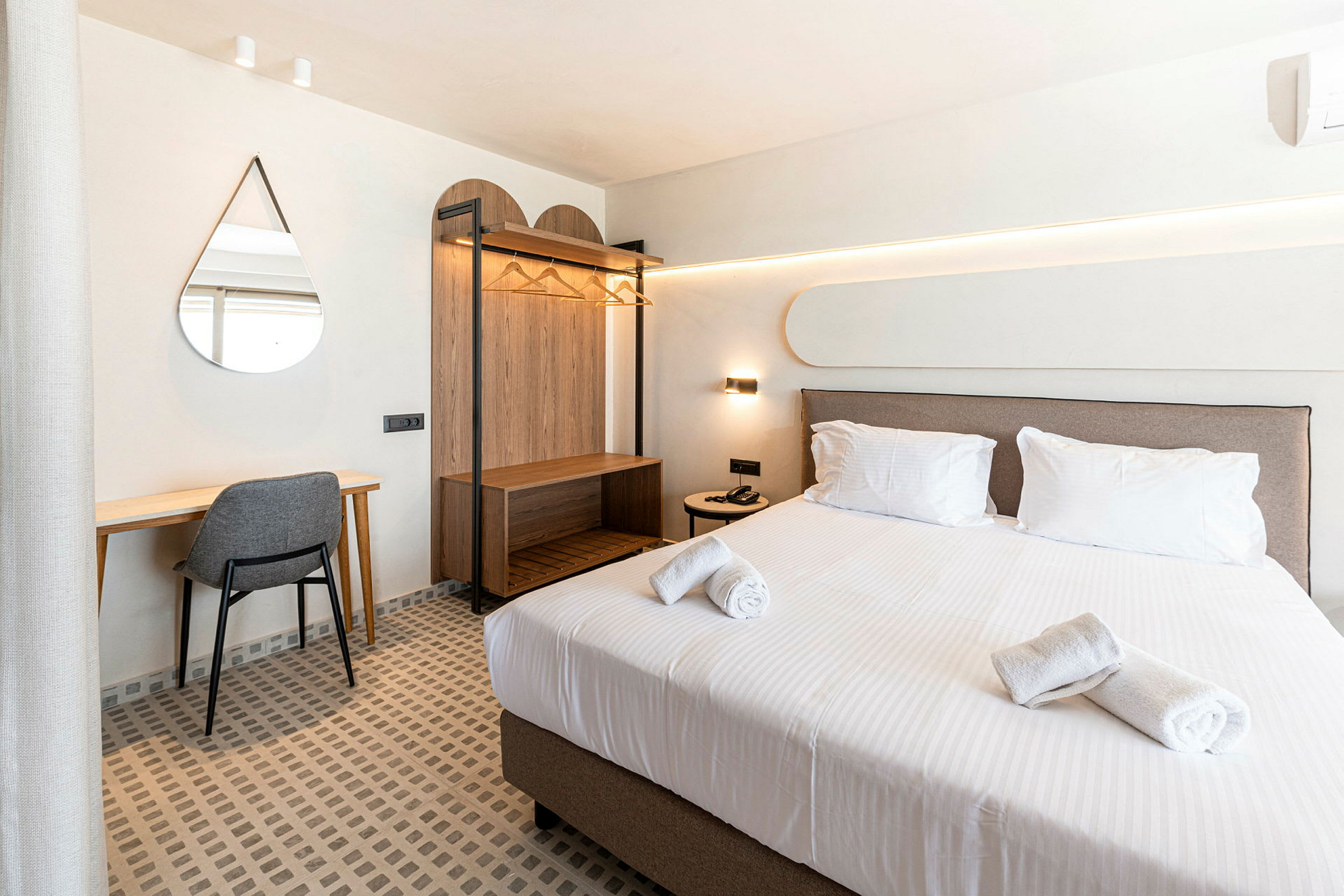 Et hotelværelse med en redt seng, skrivebord med stol, vægspejl og åben garderobe. (AI-genereret tekst)