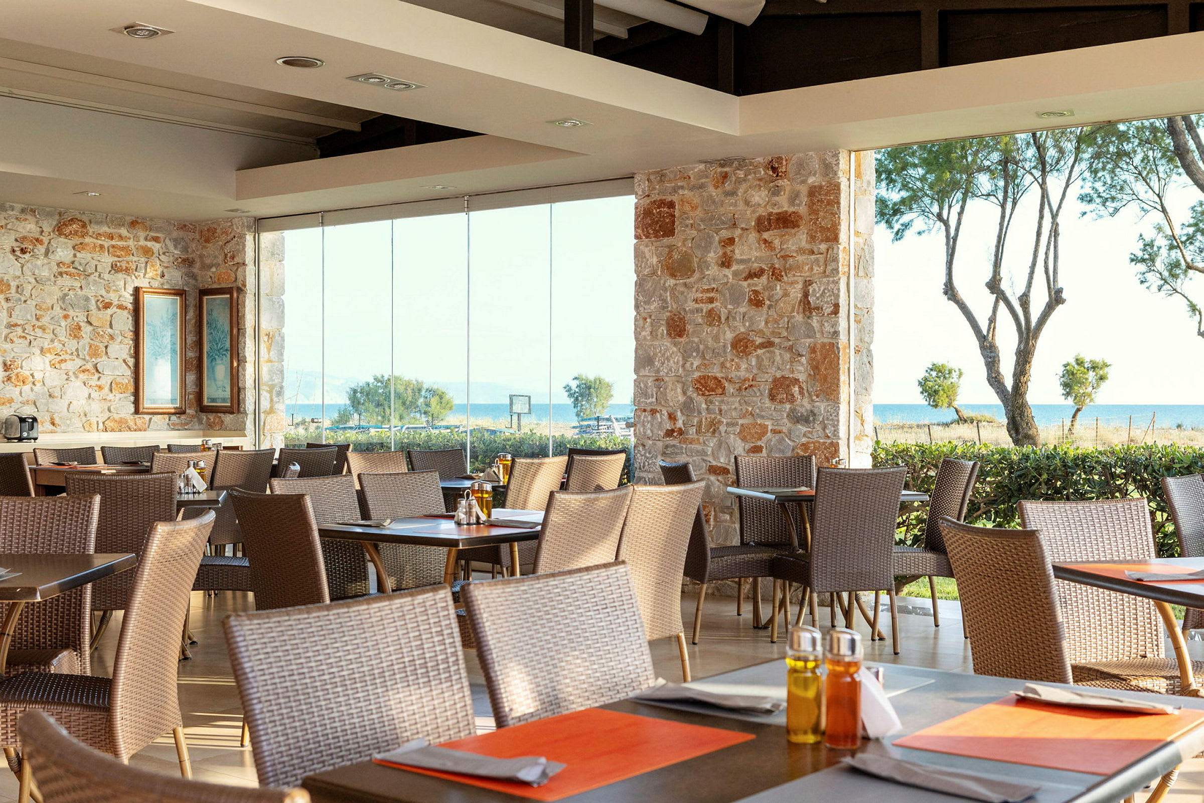 Restaurant med stenmur og store vinduer der vender ud mod havet, med flere borde og stole. (AI-genereret tekst)