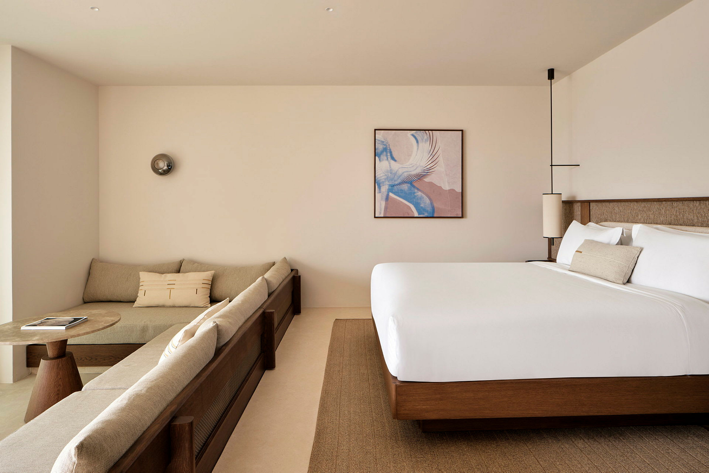 Et moderne hotelværelse med beige sofa, et rundt sofabord og en stor seng. Et billede samt dekorative lamper ses. (AI-genereret tekst)