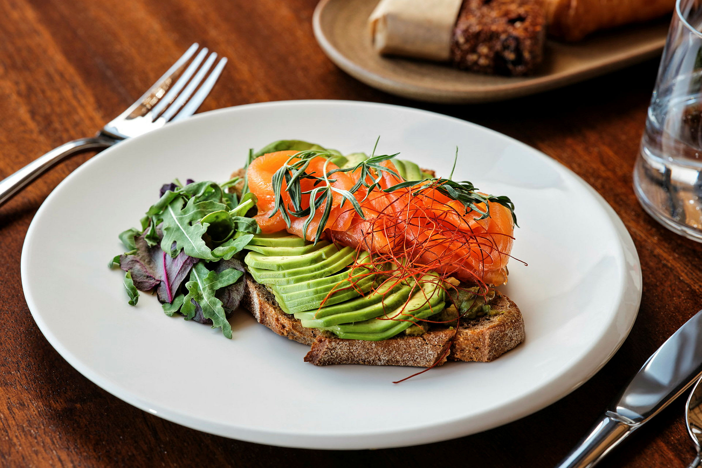 En tallerken med en ret bestående af avocado, laks og grønne blade på en skive brød. (AI-genereret tekst)