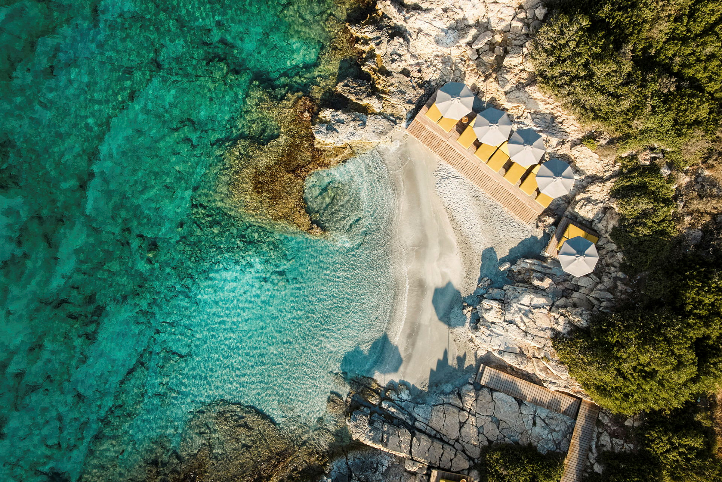 Luftfoto af en lille strand med klart turkis vand, omgivet af klipper og med parasoller ved strandkanten. (AI-genereret tekst)