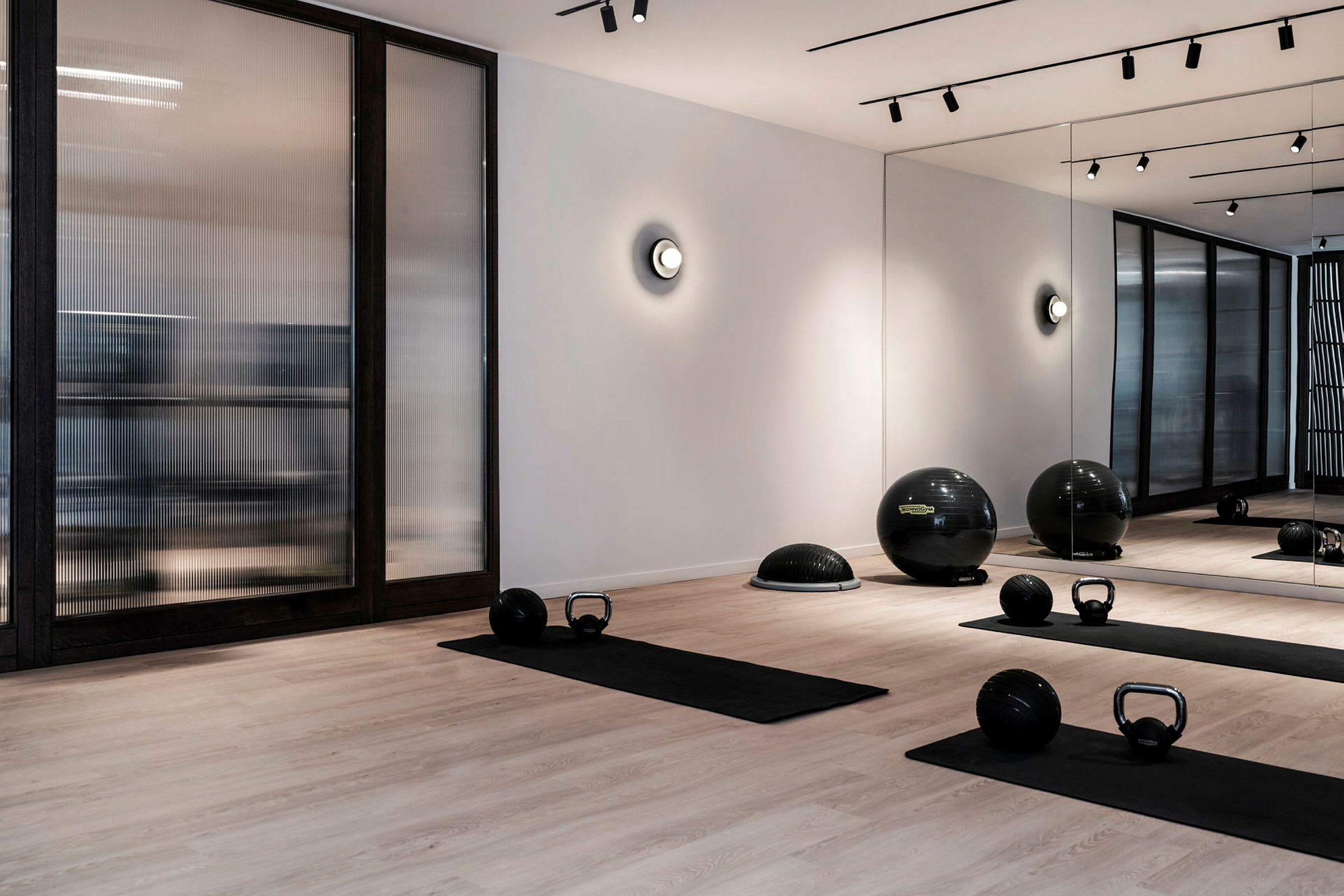 Et moderne fitnesscenter med træningsmåtter, ketlebells og fitnessbolde på et trægulv. Væggen har spejle og glaspartier. (AI-genereret tekst)