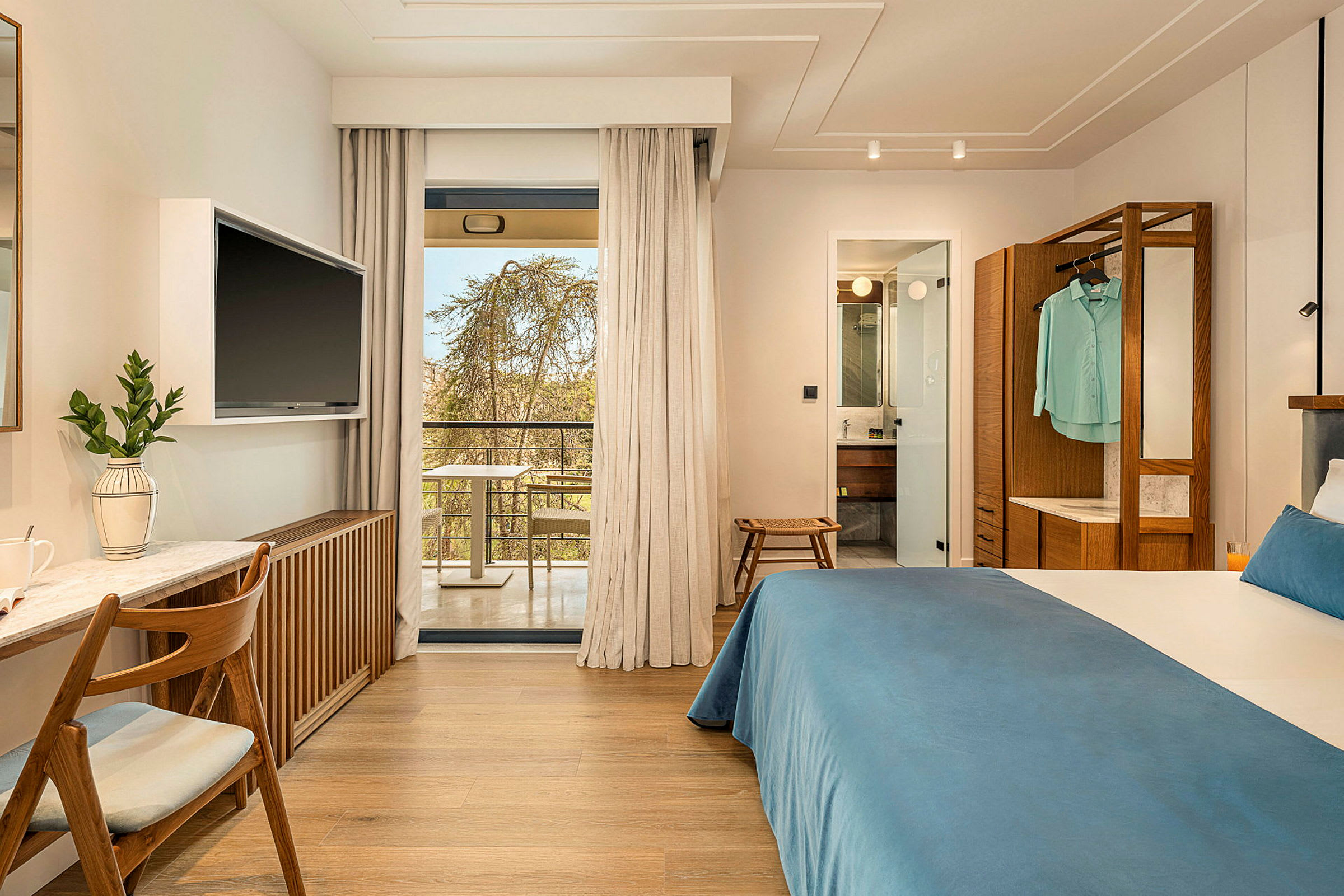 Hotelværelse med dobbeltseng, skrivebord og TV. Glasdøre fører ud til en balkon. Badeværelse synligt gennem en åben dør. (AI-genereret tekst)