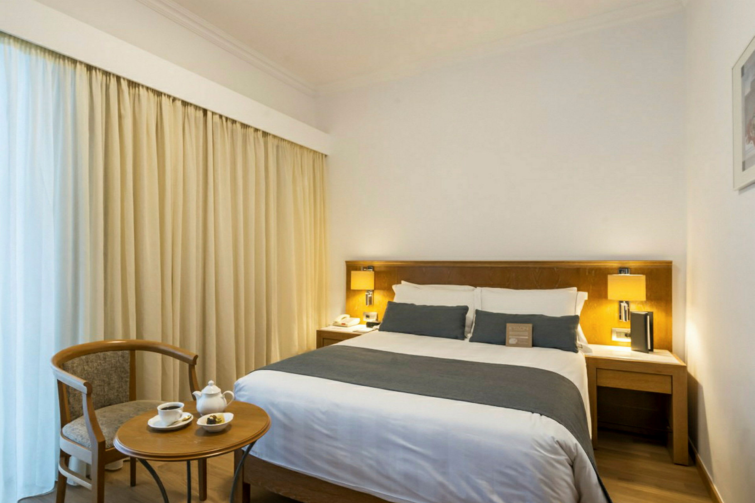 Et hotelværelse med en dobbeltseng, natborde og lamper samt et lille bord med to kopper og en stol. (AI-genereret tekst)
