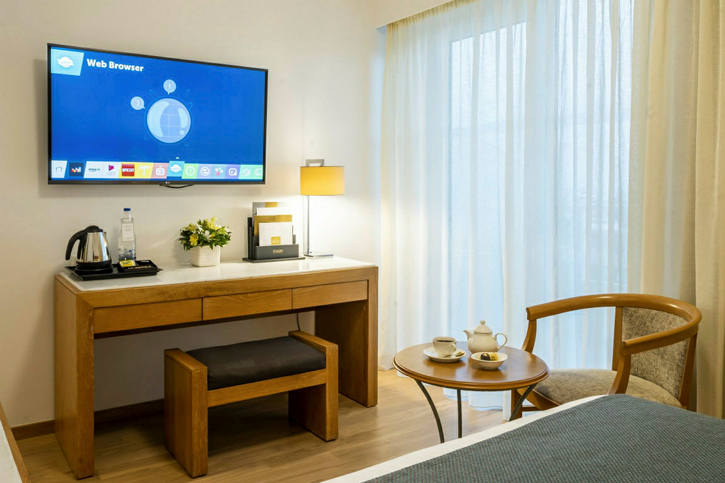 Et hotelværelse med et vægmonteret tv over et skrivebord med en stol og en elkedel på toppen. Ved siden af skrivebordet er et sidebord med tekopper og en stol. Baggrunden indeholder vinduer med lysgennemtrængende gardiner. (AI-genereret tekst)