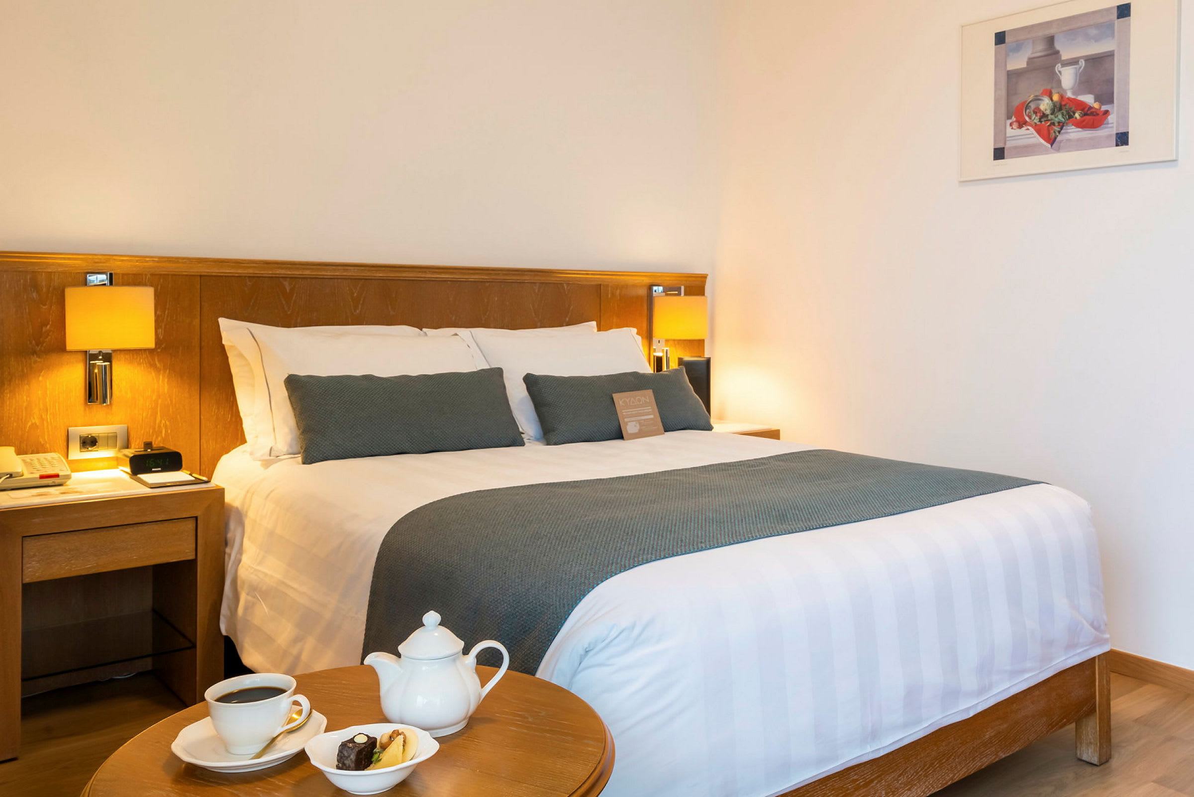 Et hotelværelse med en dobbeltseng, natborde og et bord med en kaffekop, tekande og tallerken med småkager. (AI-genereret tekst)