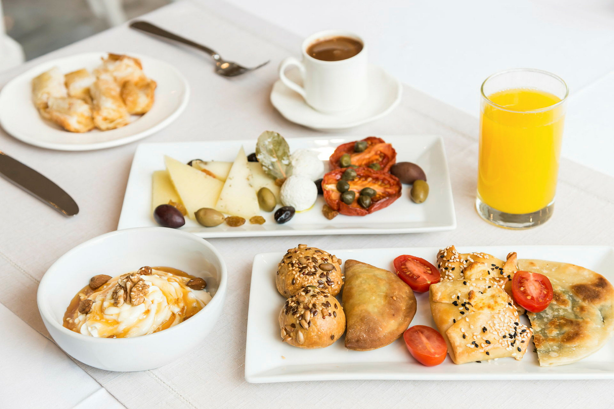 Billedet viser en morgenmadservering med forskellige ingredienser som brød, ost, soltørrede tomater, oliven og kapers. Der er også en skål med yoghurt, nogle bagværk og et glas appelsinjuice. (AI-genereret tekst)