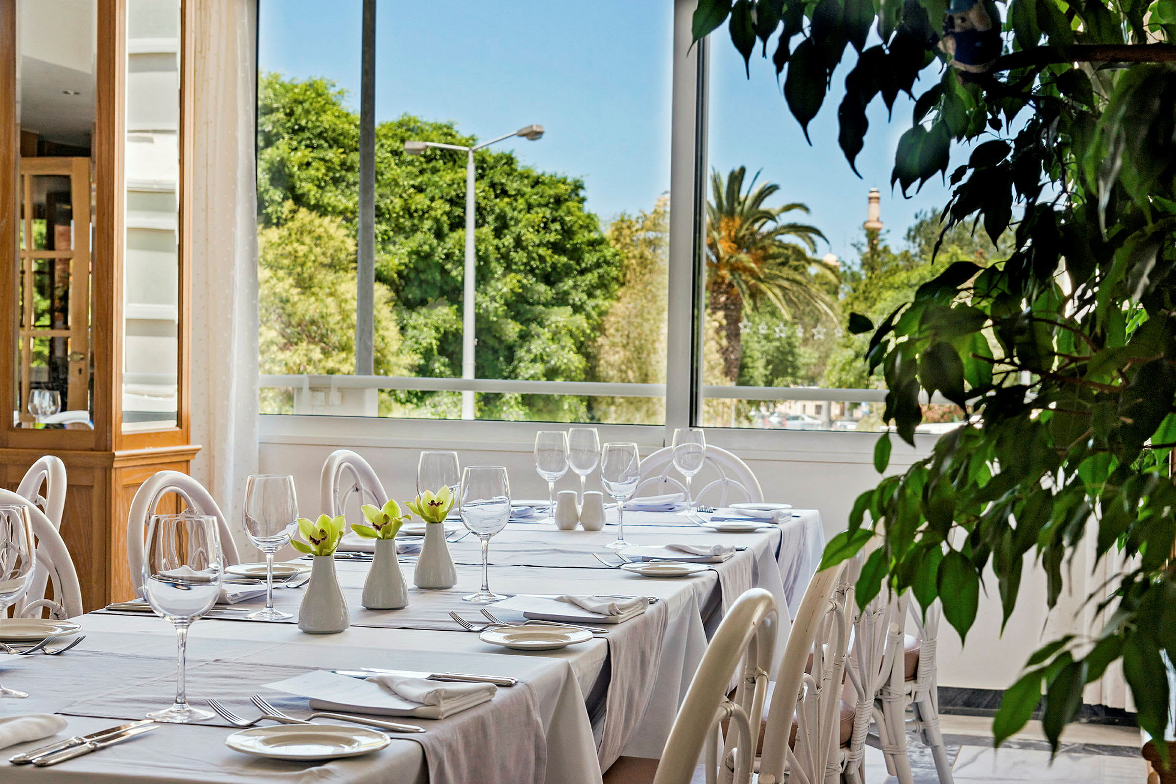 Billedet viser et opdækket bord i en restaurant med hvidt porcelæn og glas. I baggrunden er der store vinduer med udsigt til frodig natur. (AI-genereret tekst)