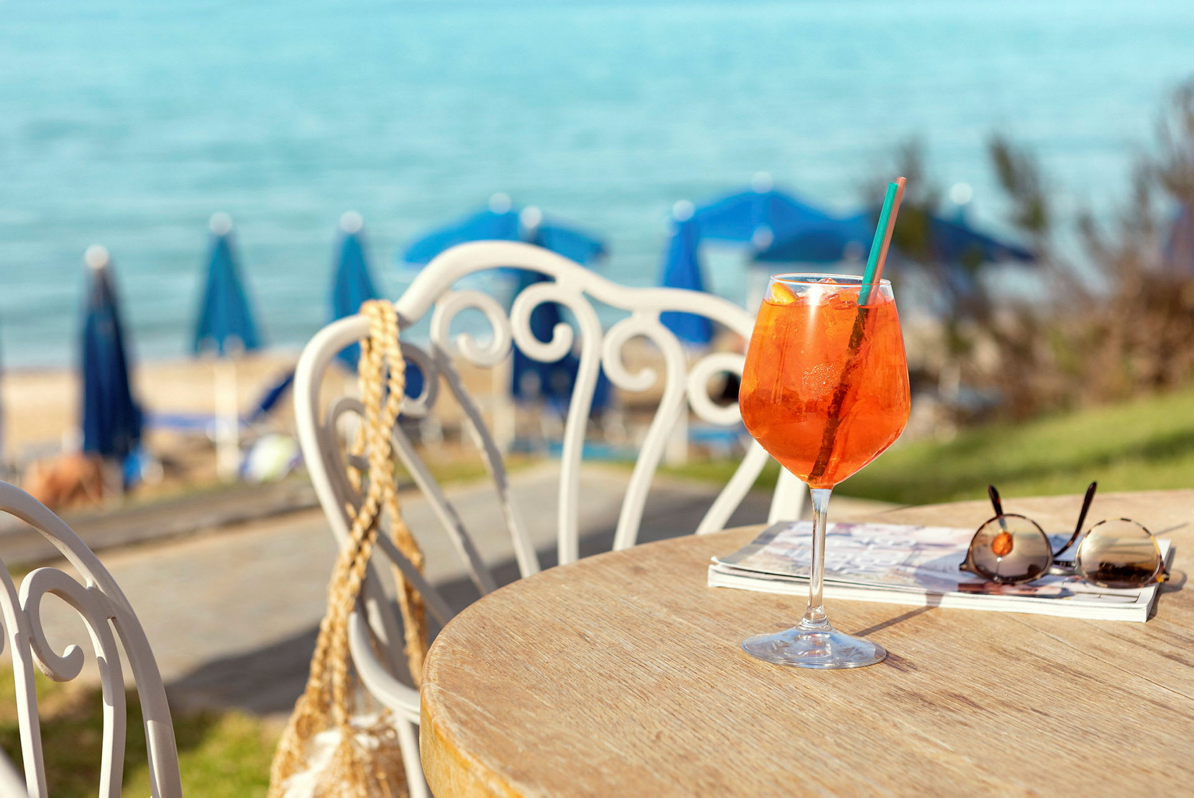 Et bord med et glas appelsindrik, solbriller og en bog. I baggrunden ses en hav og blå parasoller på stranden. (AI-genereret tekst)