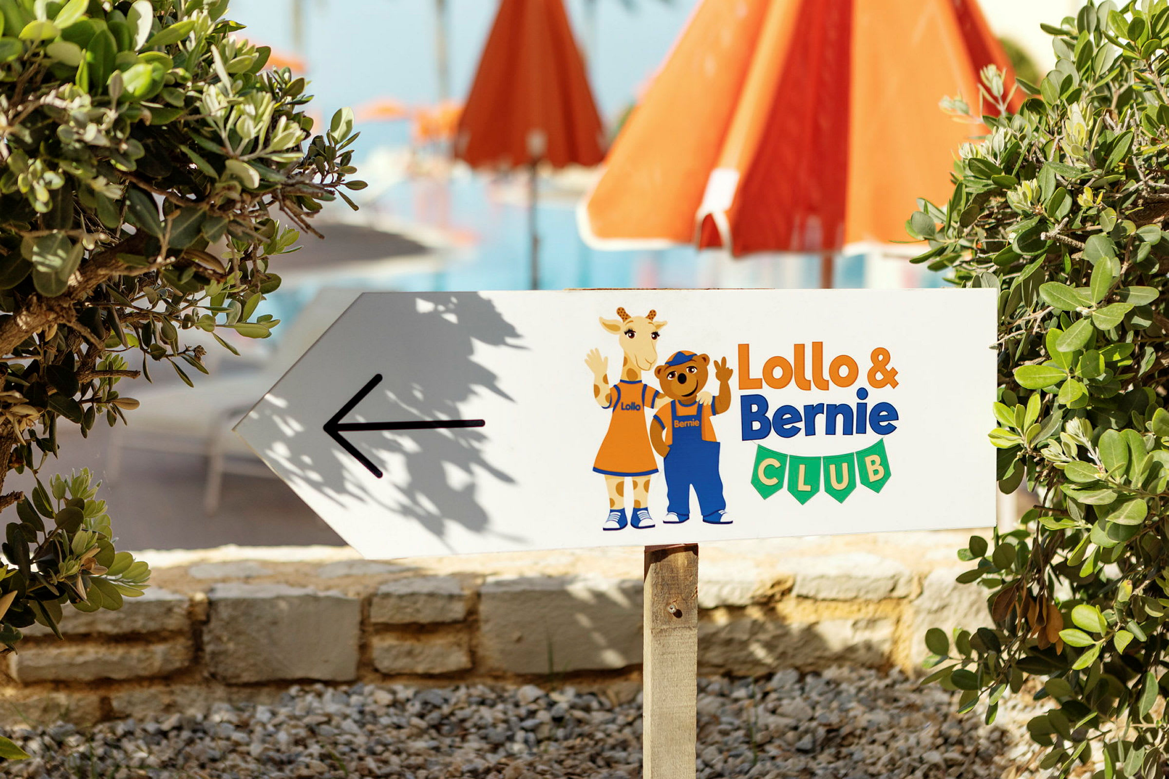Skilt med pil og teksten 'Lollo & Bernie Club', med illustrationer af Lollo og Bernie. Baggrund med grønt og røde parasoller. (AI-genereret tekst)