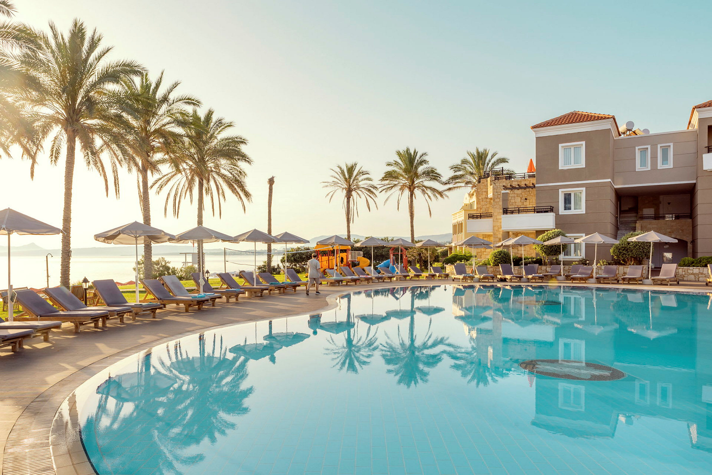 Udendørs swimmingpool med liggestole og parasoller ved hotelområde, omgivet af palmer og med udsigt over havet. (AI-genereret tekst)