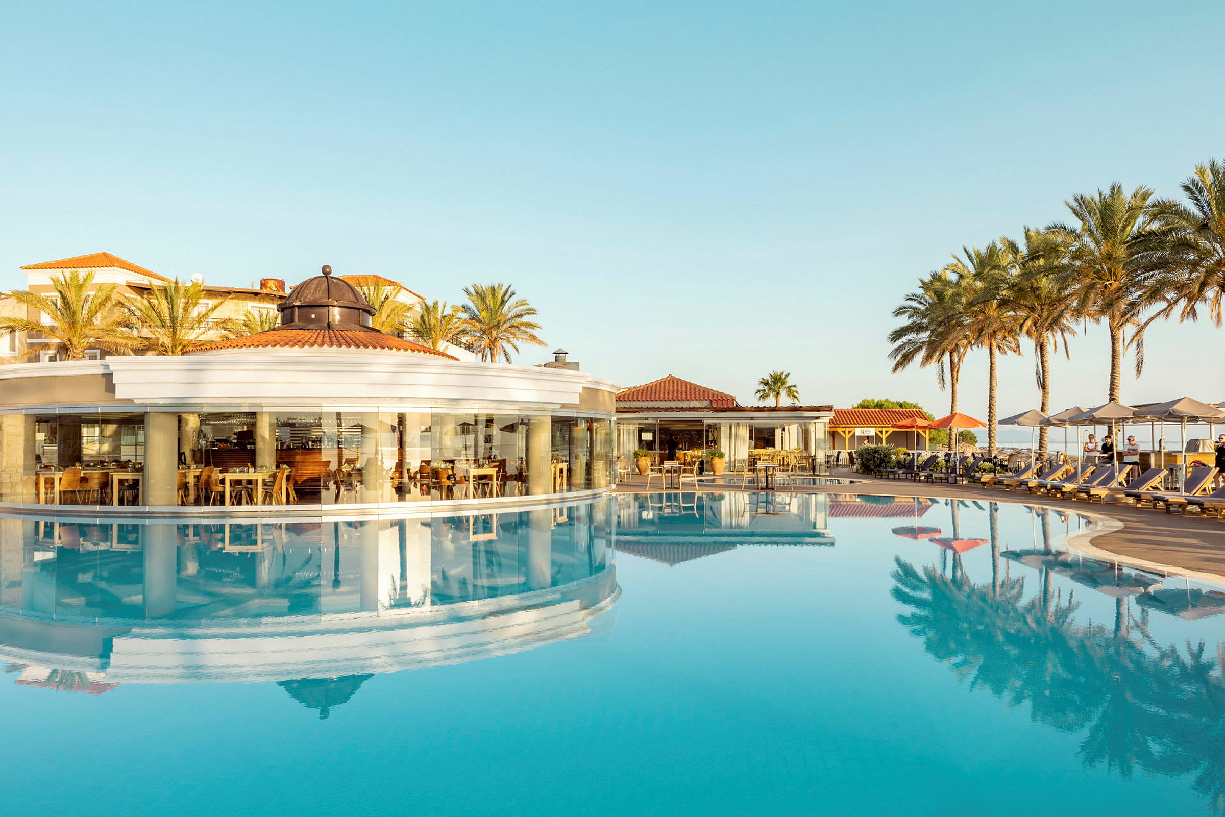 En restaurant med panoramaglasvægge ved en pool, omgivet af palmer. (AI-genereret tekst)