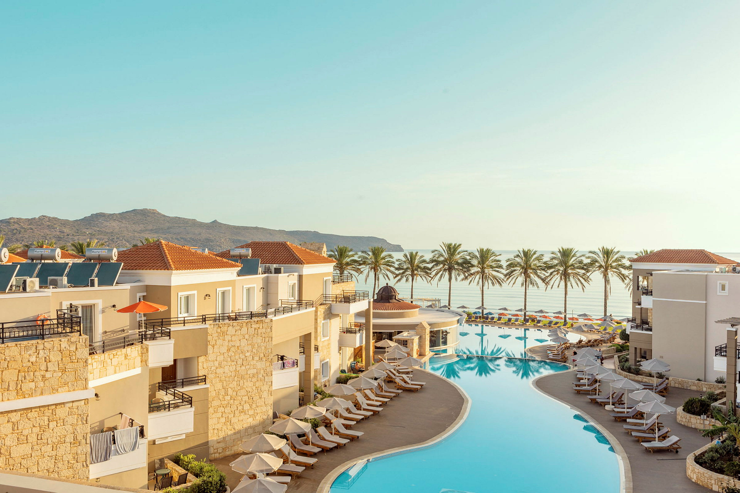 Oversigt over et resort med pool og liggestole, med havudsigt og palmer i baggrunden. (AI-genereret tekst)