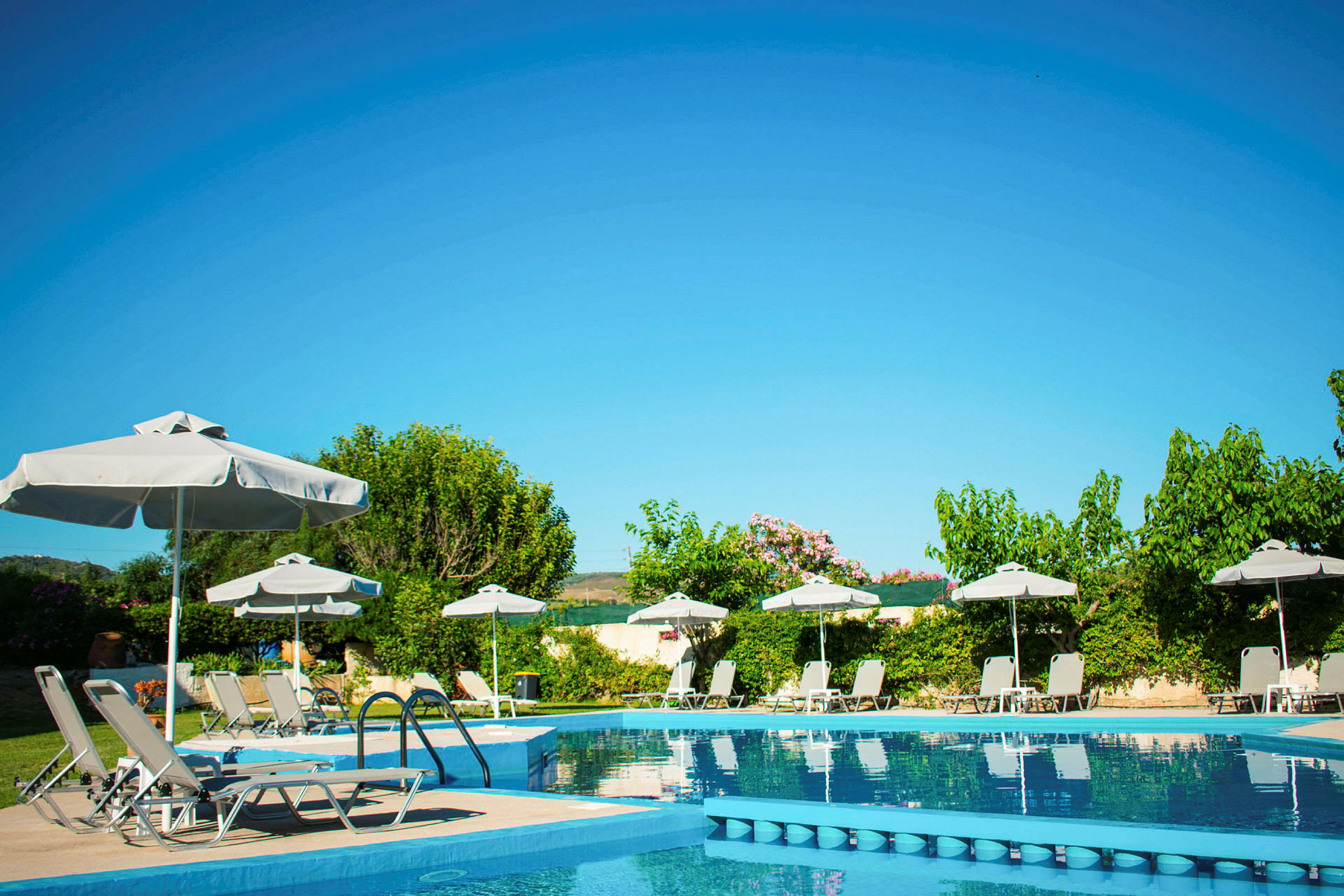 En swimmingpool med liggestole, parasoller og frodige omgivelser under en klar blå himmel. (AI-genereret tekst)