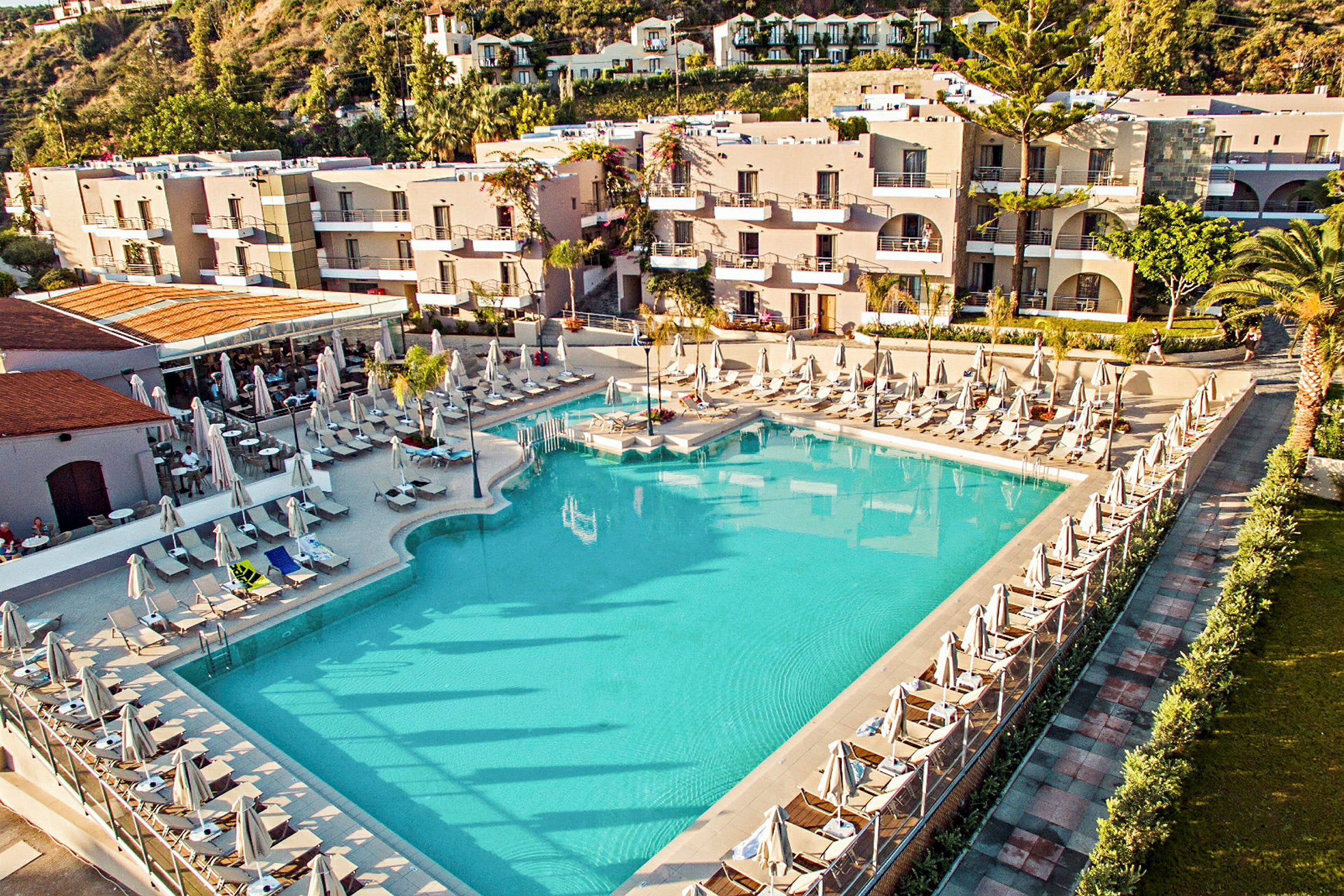 En stor blå swimmingpool omgivet af liggestole og parasoller foran en hotelbygning. (AI-genereret tekst)