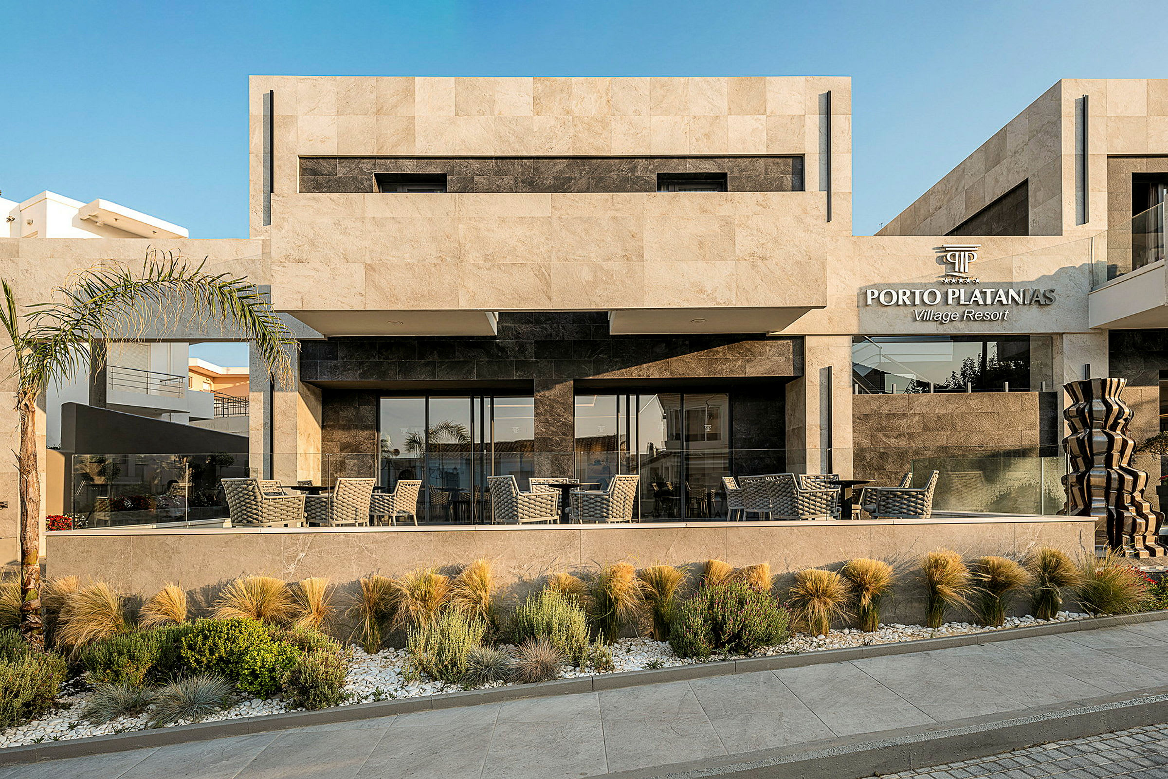 Udvendig af Porto Platanias Village Resort bygning med moderne design og udendørs servering. (AI-genereret tekst)