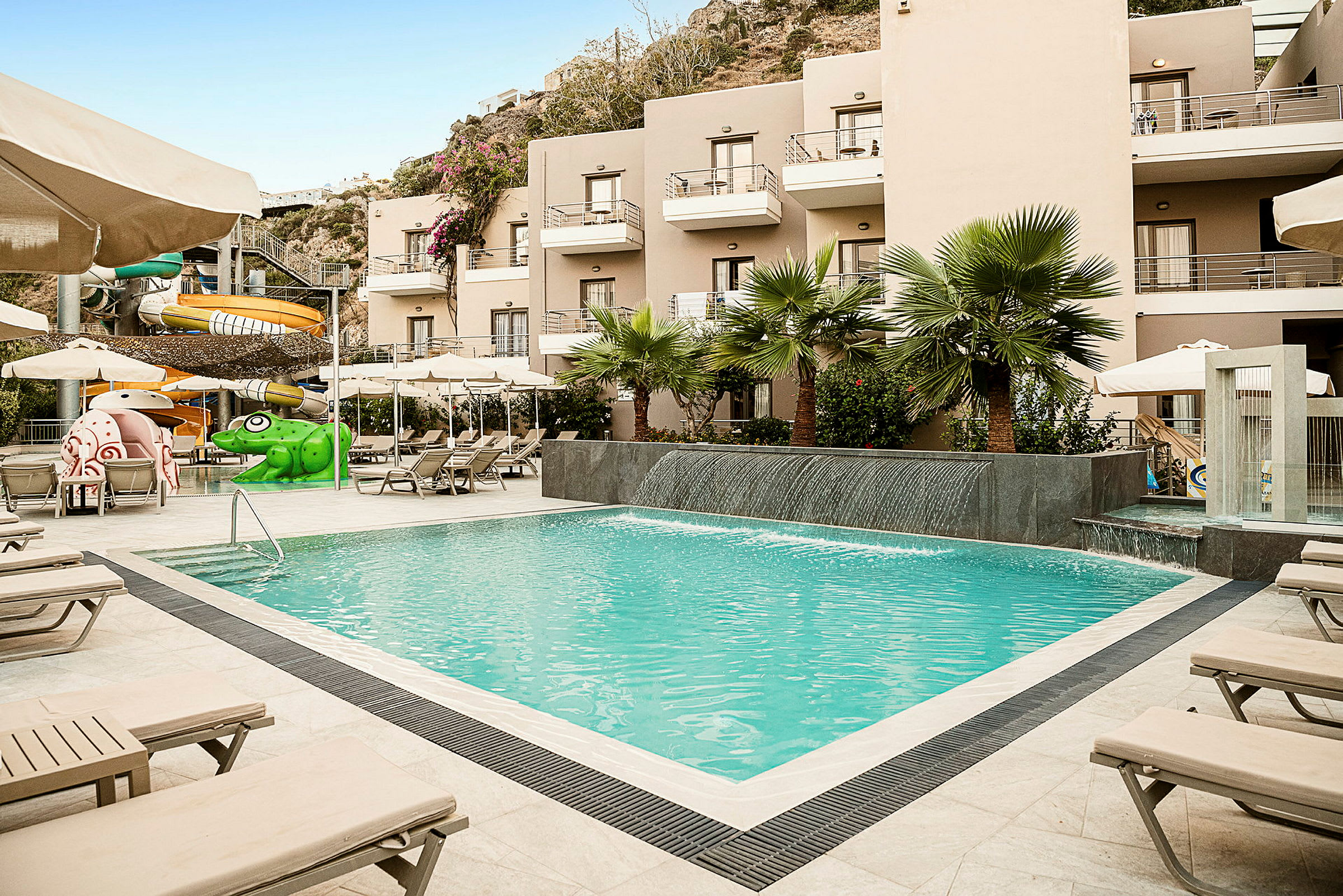En udendørs pool ved siden af et hotel med liggestole, vandrutsjebaner og vegetation. (AI-genereret tekst)