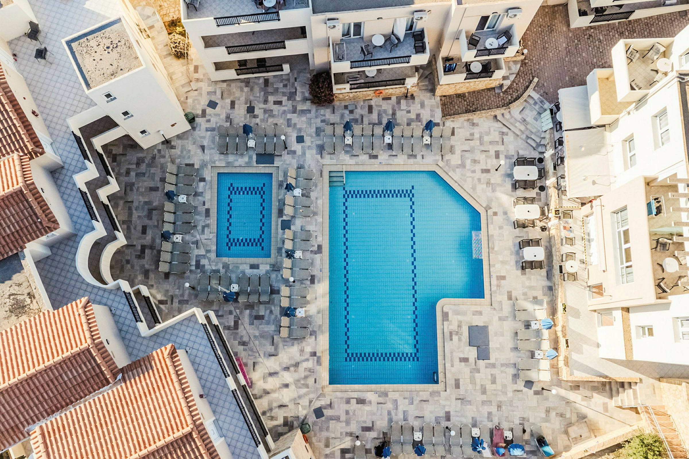 Luftfoto af et hotelområde med to rektangulære pools omgivet af solsenge og bygninger. (AI-genereret tekst)