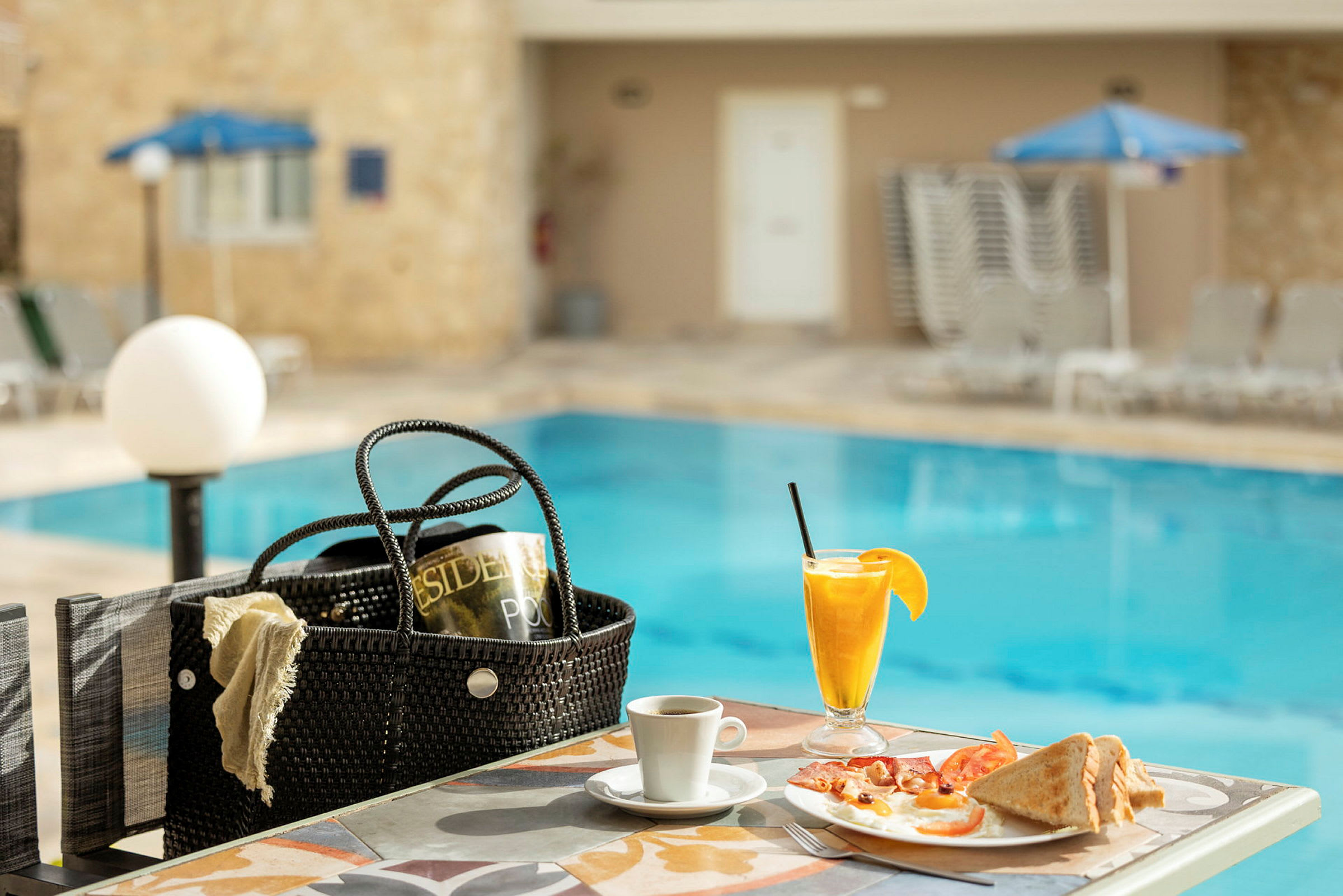 Morgenmad serveret ved et bord tæt på en swimmingpool, inklusive appelsinjuice, kaffe og en tallerken med brød og æg. (AI-genereret tekst)